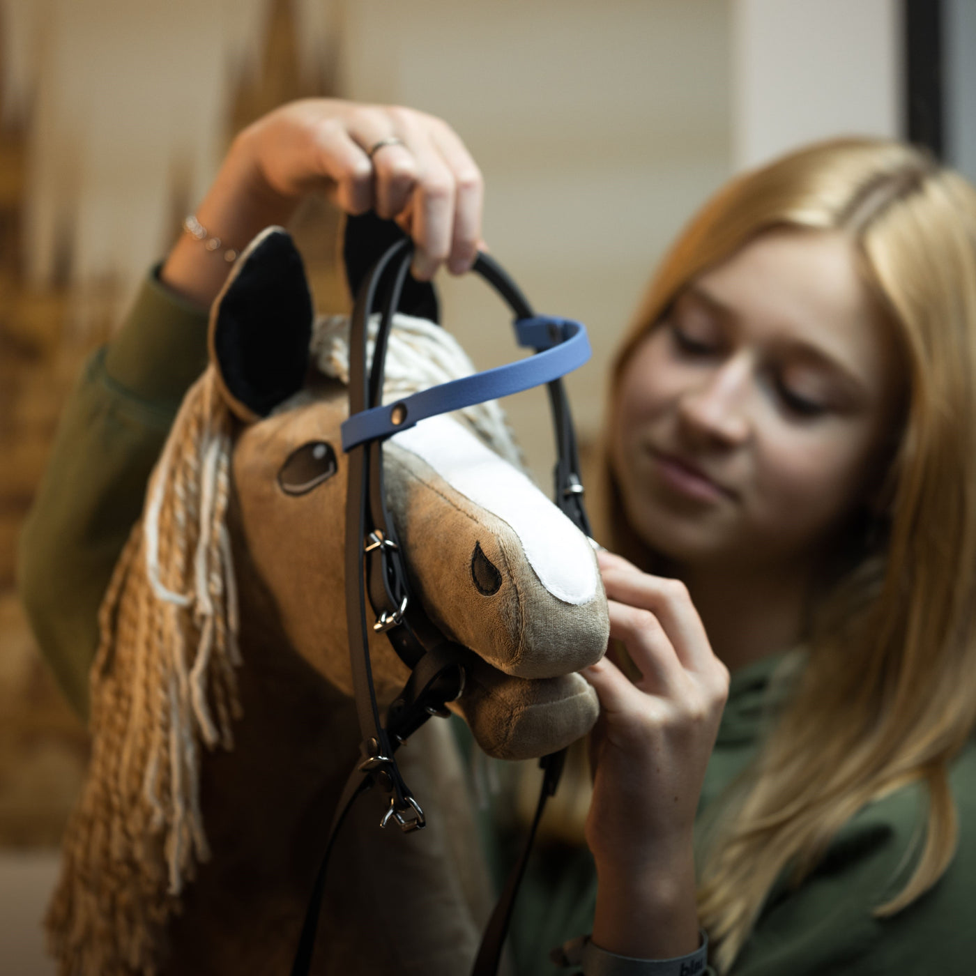 Dívka s blond vlasy nasazuje uzdečku svému Hobby Horse a soustředěně upravuje řemínky – zachycuje ducha hobby horsing.