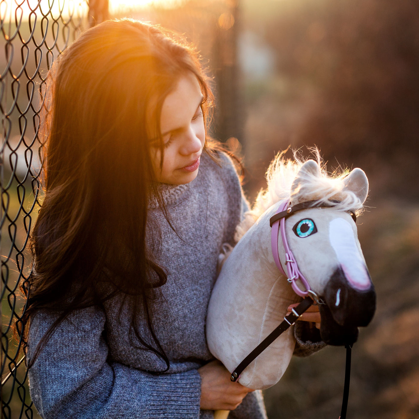 Mladá dívka s dlouhými tmavými vlasy v šedém svetru stojí při západu slunce u drátěného plotu a drží svého oblíbeného hobby horse – odkaz na hravý svět hobby horsing – jeho bílá hříva a růžová uzdečka zachycují zlaté světlo.