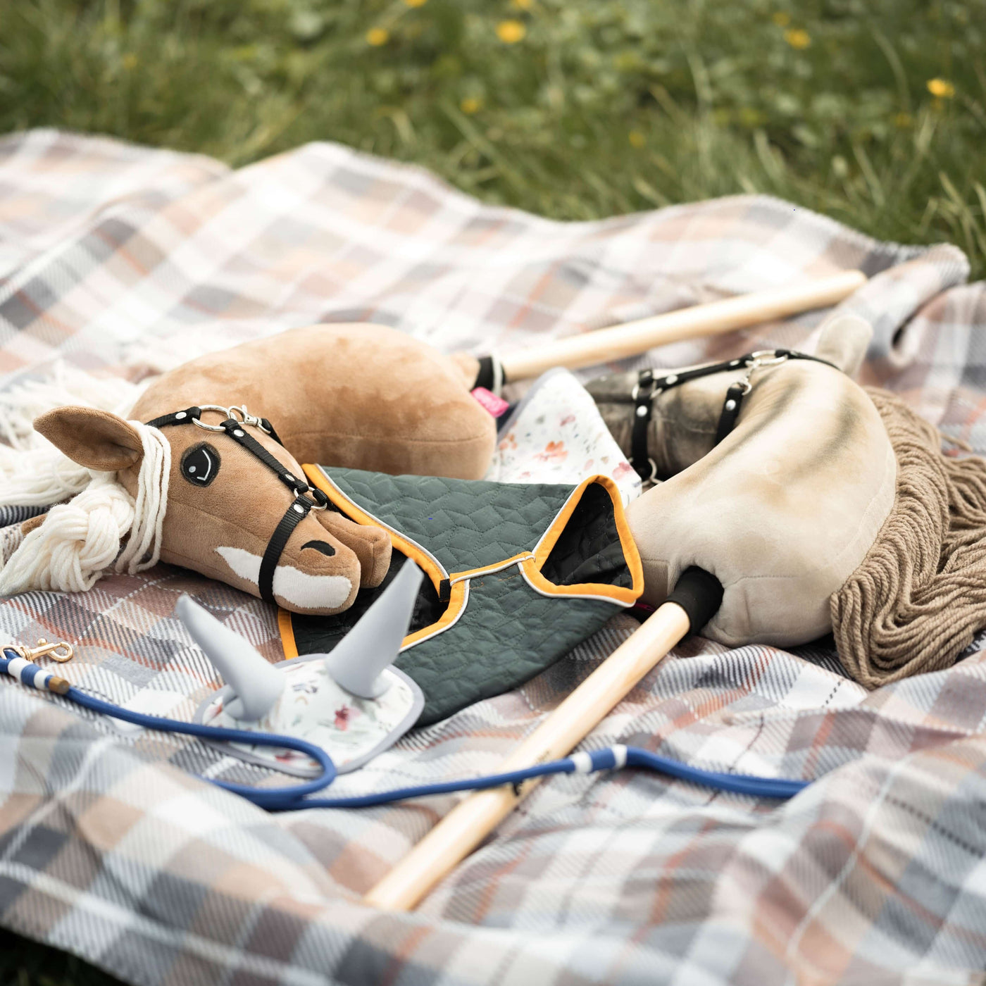 Dvě hračky Hobby Horse s látkovými hlavami, otěžemi a dřevěnými tyčemi leží na kostkované dece rozprostřené na trávě vedle malé šedé hračky připomínající koňskou hlavu a zelené prošívané látky – ideální pro nápaditá dobrodružství s Hobby Horsing.