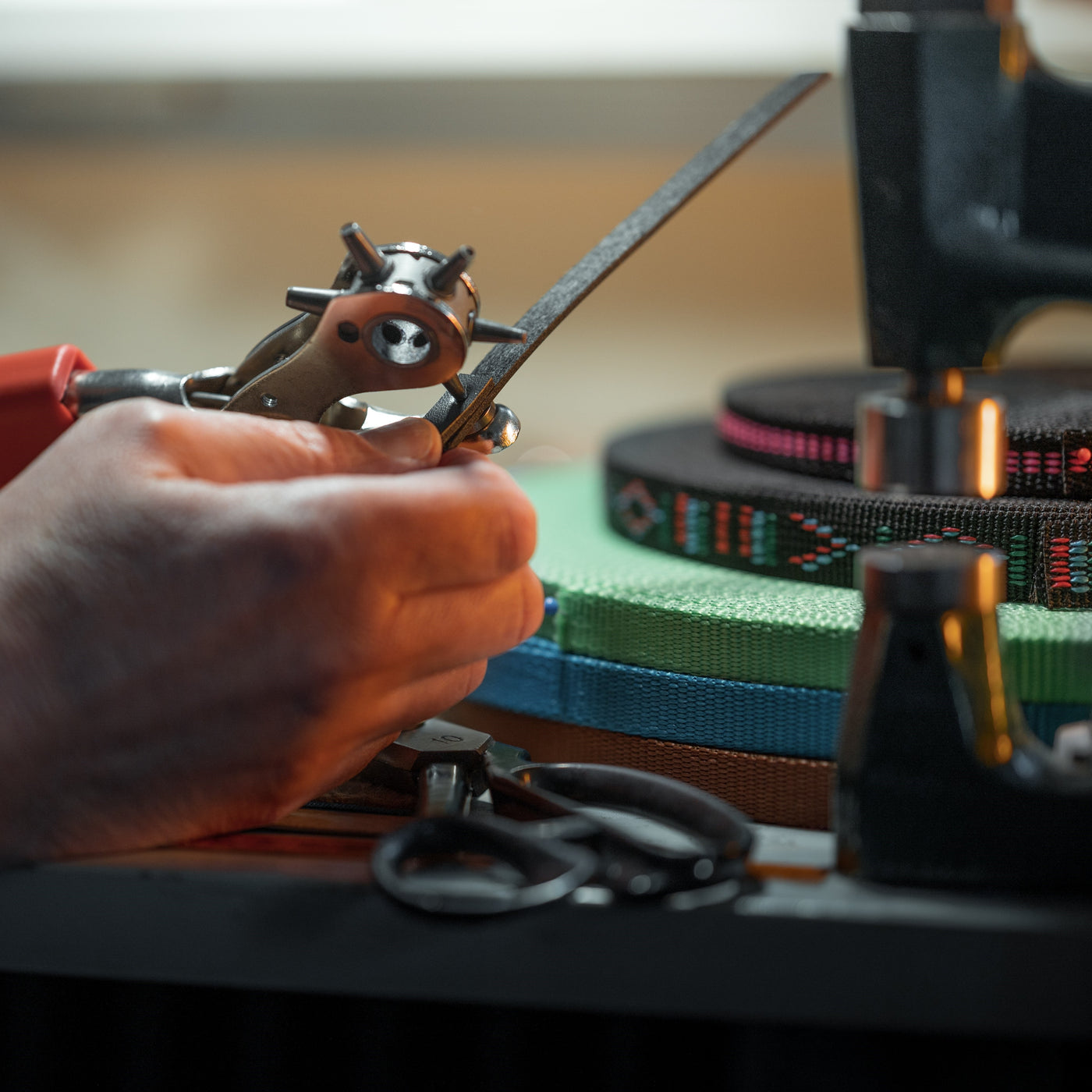 Detail osoby používající děrovací nástroj na pruh materiálu pro projekt Hobby Horse, přičemž v pozadí na pracovním stole jsou vidět barevné tkané popruhy a nůžky.