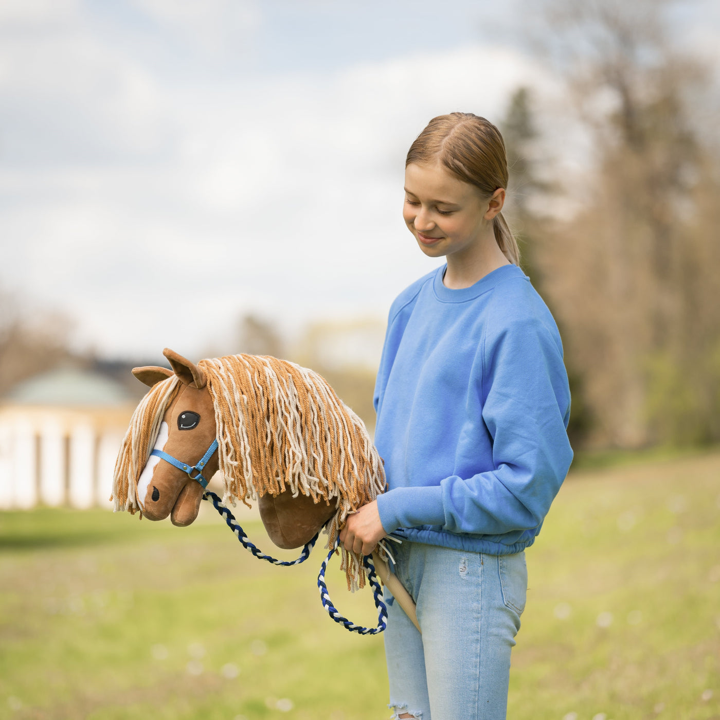 Mladý člověk v modré mikině a džínách stojí venku a s úsměvem se dívá na hnědého hobby horse s provazovou uzdečkou, užívá si zábavu hobby horsing na travnatém trávníku se stromy v pozadí.