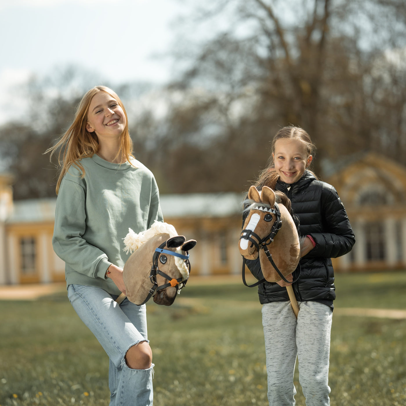 Dvě usmívající se dívky stojí venku a drží své hobby horse. Jedna má na sobě světle zelený svetr a roztrhané džíny, druhá černou bundu a šedé kalhoty. Hobby horsing jim přináší radost, když pózují poblíž velké budovy a stromů v pozadí.