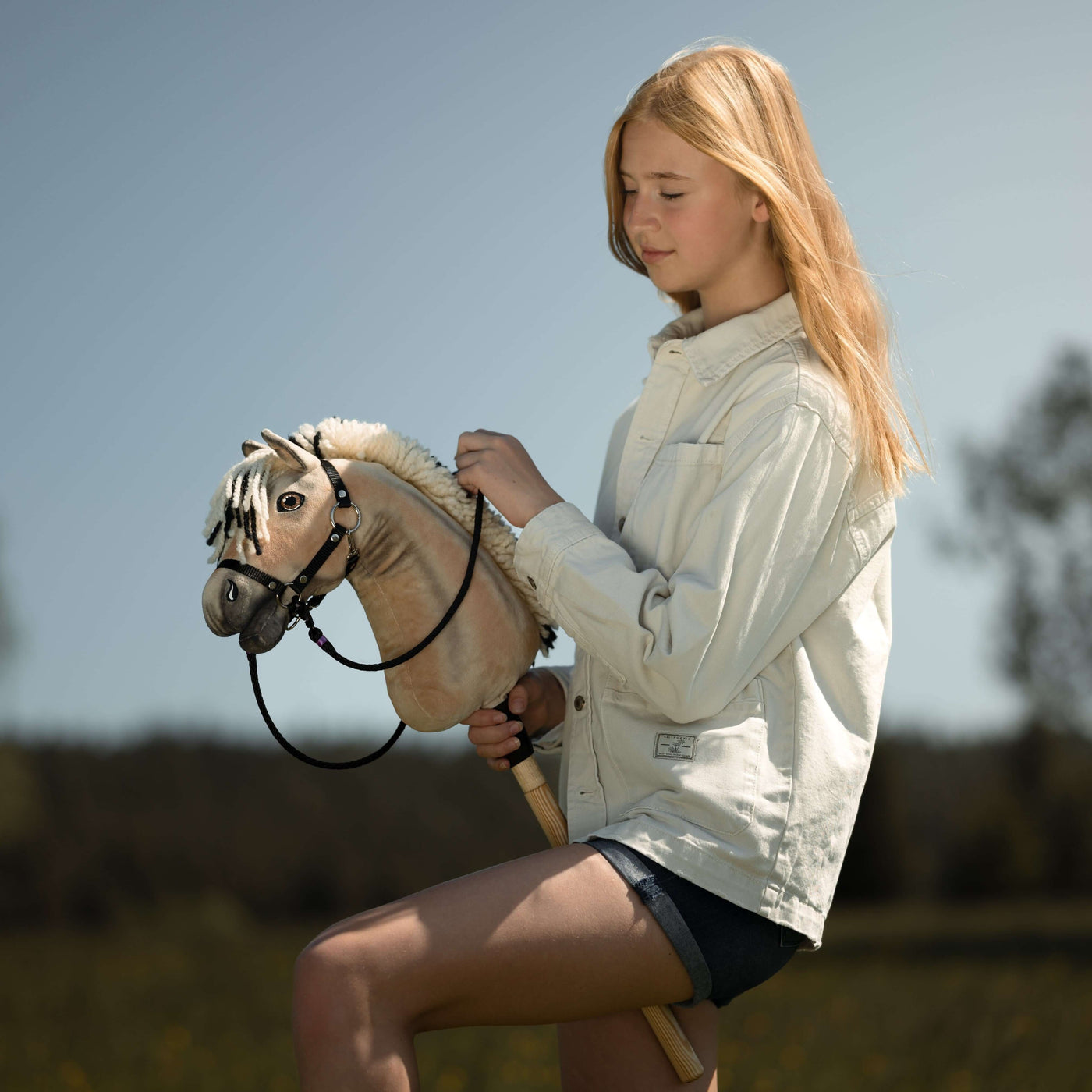 Mladá dívka s dlouhými blond vlasy, v lehké bundě a kraťasech, si za slunečného dne venku užívá Hobby Horsing a drží svůj Hobby Horse mezi stromy a na rozostřeném přírodním pozadí.
