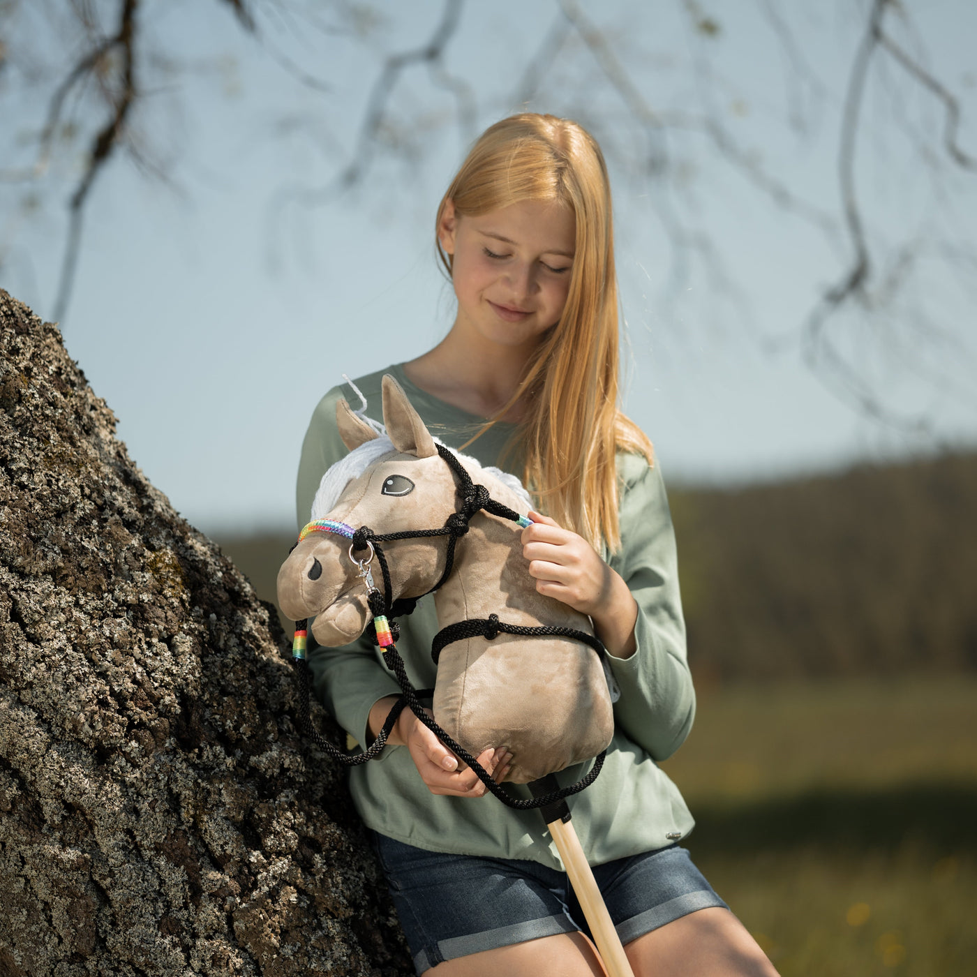 Mladá dívka s dlouhými blond vlasy, v zeleném topu a džínových kraťasech, sedí venku u stromu a s úsměvem se dívá na svého Hobby Horse s uzdečkou a barevnými ozdobami – zachycuje radostného ducha Hobby Horsing v přírodním prostředí.