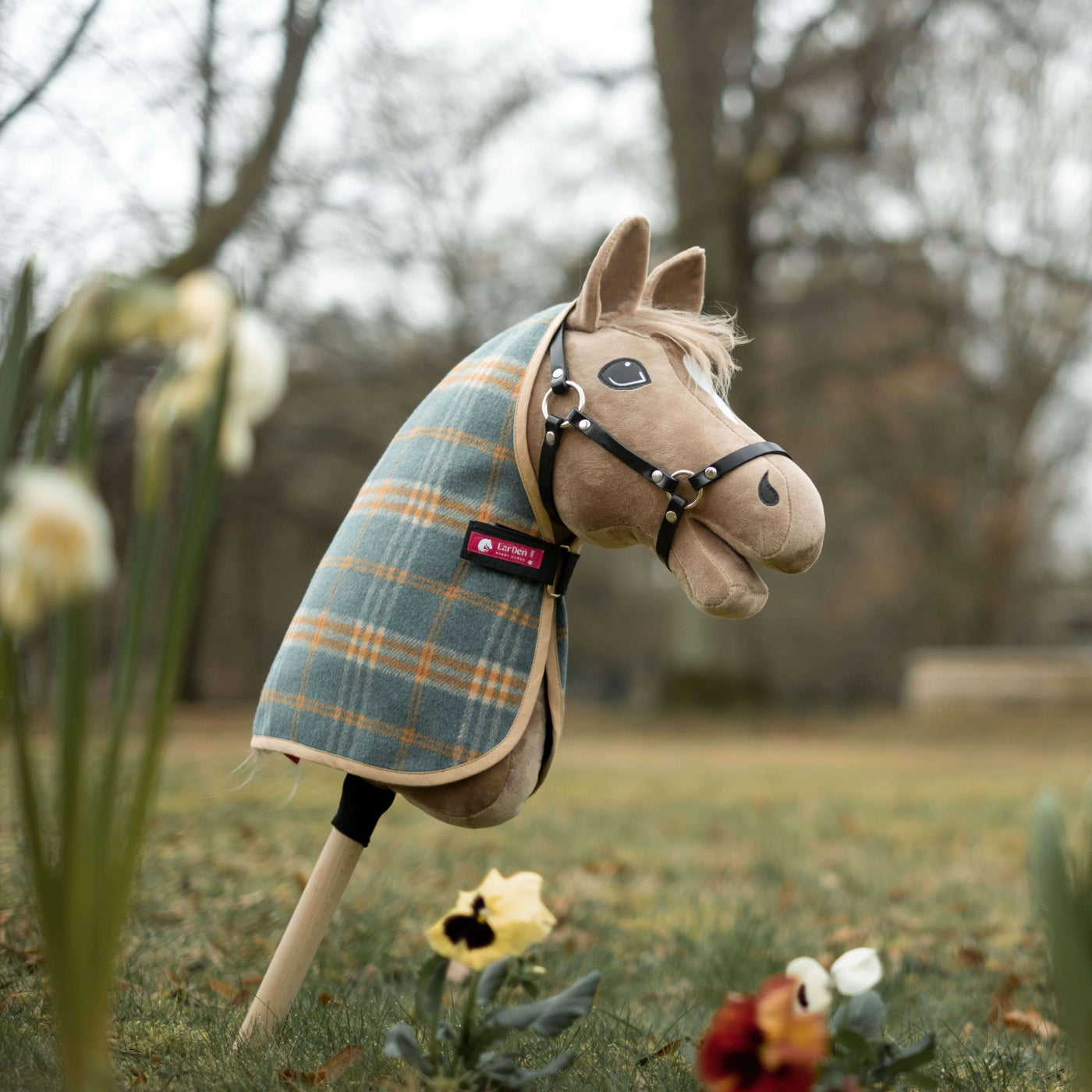 Plyšový Hobby Horse s kostkovanou dekou a uzdečkou stojí vzpřímeně na tyči v travnatém venkovním prostředí, ideální pro Hobby Horsing dobrodružství mezi jarními květinami a stromy v pozadí.