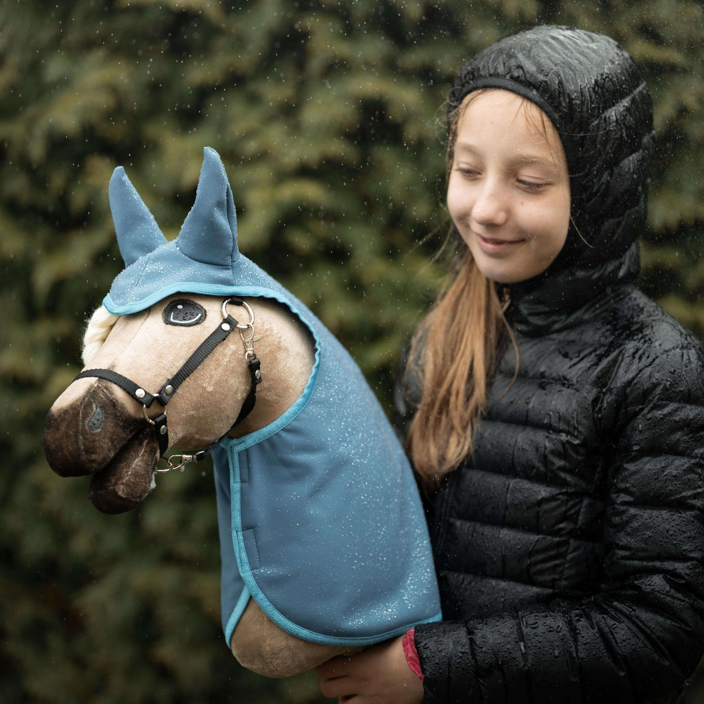 Usměvavá dívka v černé bundě s kapucí stojí venku v dešti a drží svého oblíbeného plyšového Hobby Horse oblečeného v modré, zatímco v pozadí je rozostřená zelená zeleň – dokonalý okamžik pro každého nadšence do Hobby Horsing.
