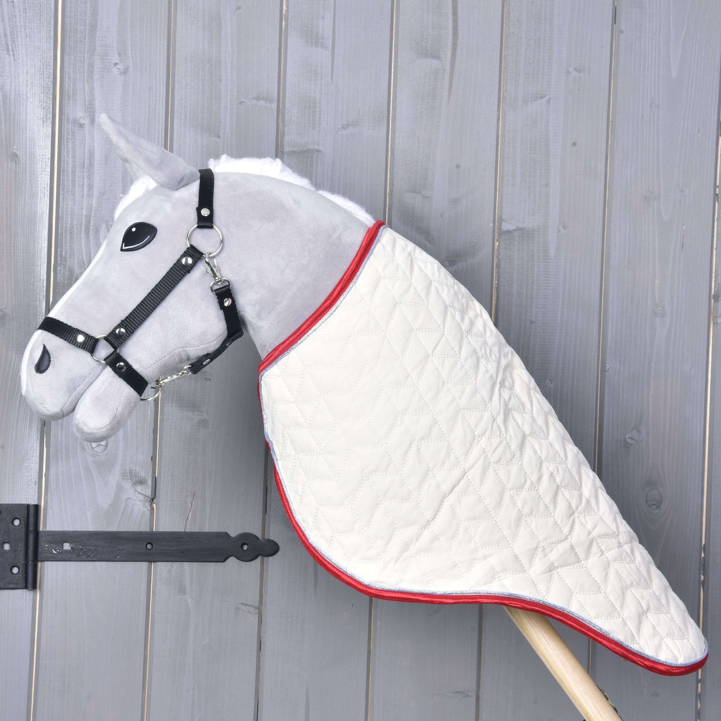 Hobby Horse LarDen Převozní deka Italy na šedém plyšovém Hobby Horse s černou uzdečkou a prošívanou bílou dekou, na dřevěné tyči před šedou dřevěnou stěnou.