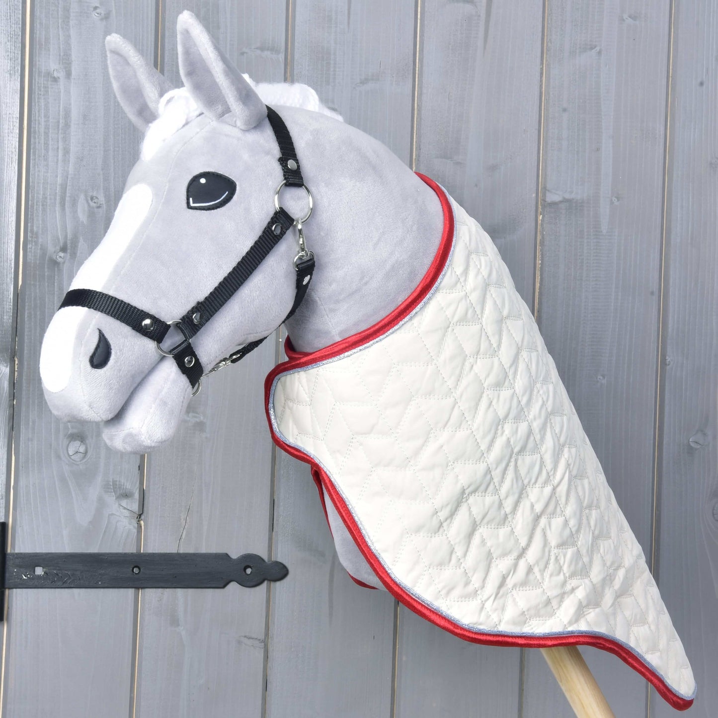 Převozní deka Italy od Hobby Horse LarDen na šedém plyšovém Hobby Horse s černou uzdečkou; prošívaná bílá deka s červeným lemováním, na dřevěné tyči před světle šedým dřevěným pozadím.