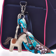 Detail tmavě modré tašky s růžovým lemováním, na které je připevněný Přívěšek na klíče Horse Grey od Hobby Horse LarDen a barevný vzorovaný šátek uvázaný do mašle na jednom z kovových kroužků tašky.