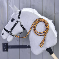 Hobby Horse - pletené vodítko Pomello od Hobby Horse LarDen na plyšovém šedém Hobby Horse s bílou hřívou, černou uzdečkou a oranžovo-černým pleteným vodítkem s karabinou, připevněném na dřevěné stěně.