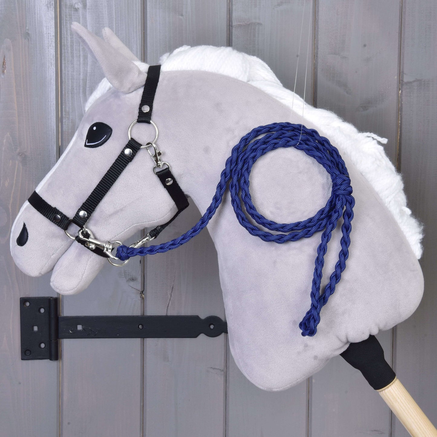 Hobby Horse - pletené vodítko Royal Blue od Hobby Horse LarDen na šedém plyšovém Hobby Horse s bílou hřívou, černou uzdečkou a královsky modrým ručně pleteným vodítkem, připevněném na dřevěné stěně s tyčí zajištěnou černým kovovým držákem.