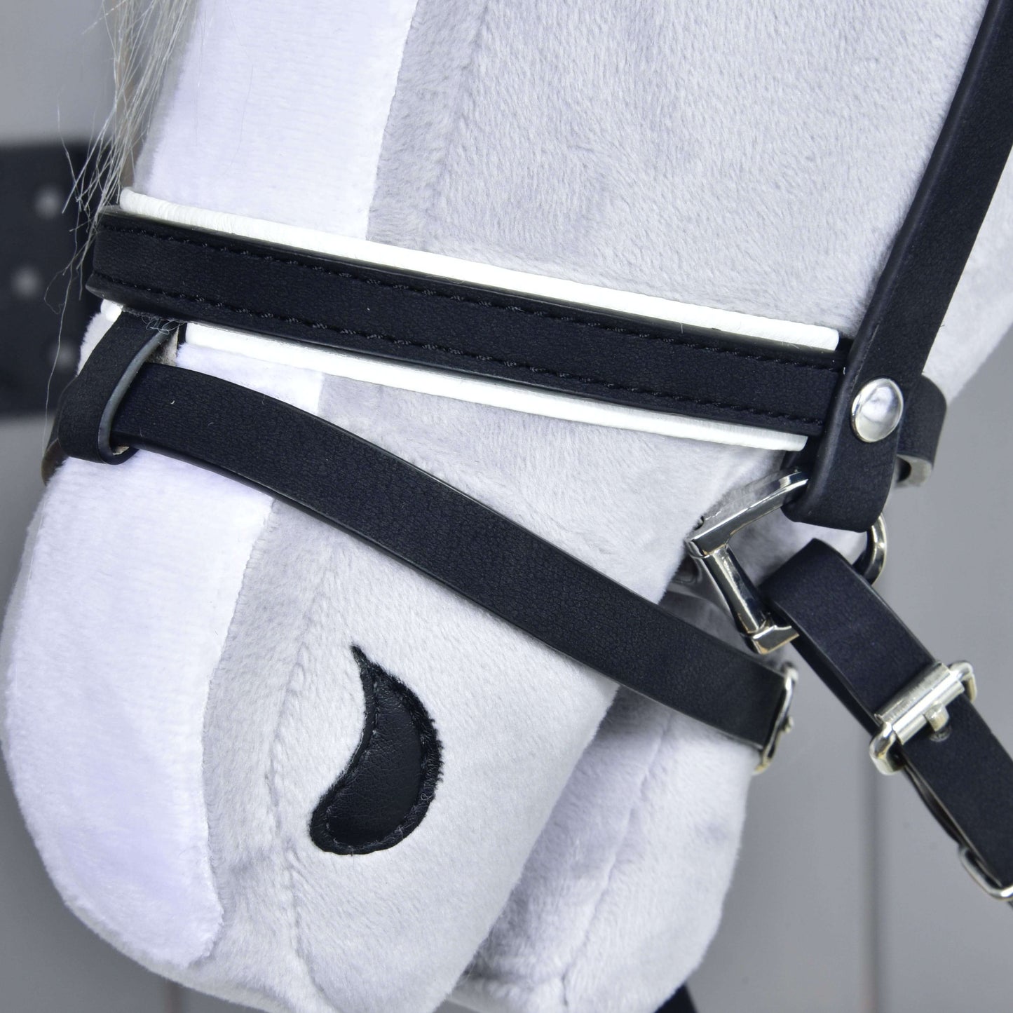 Detail hlavy šedého plyšového koně od Hobby Horse LarDen s kompletní uzdečkou De Luxe se 2 čelenkami. Vyšívané detaily, jako černá nozdra ve tvaru půlměsíce, vynikají na světlém pozadí.