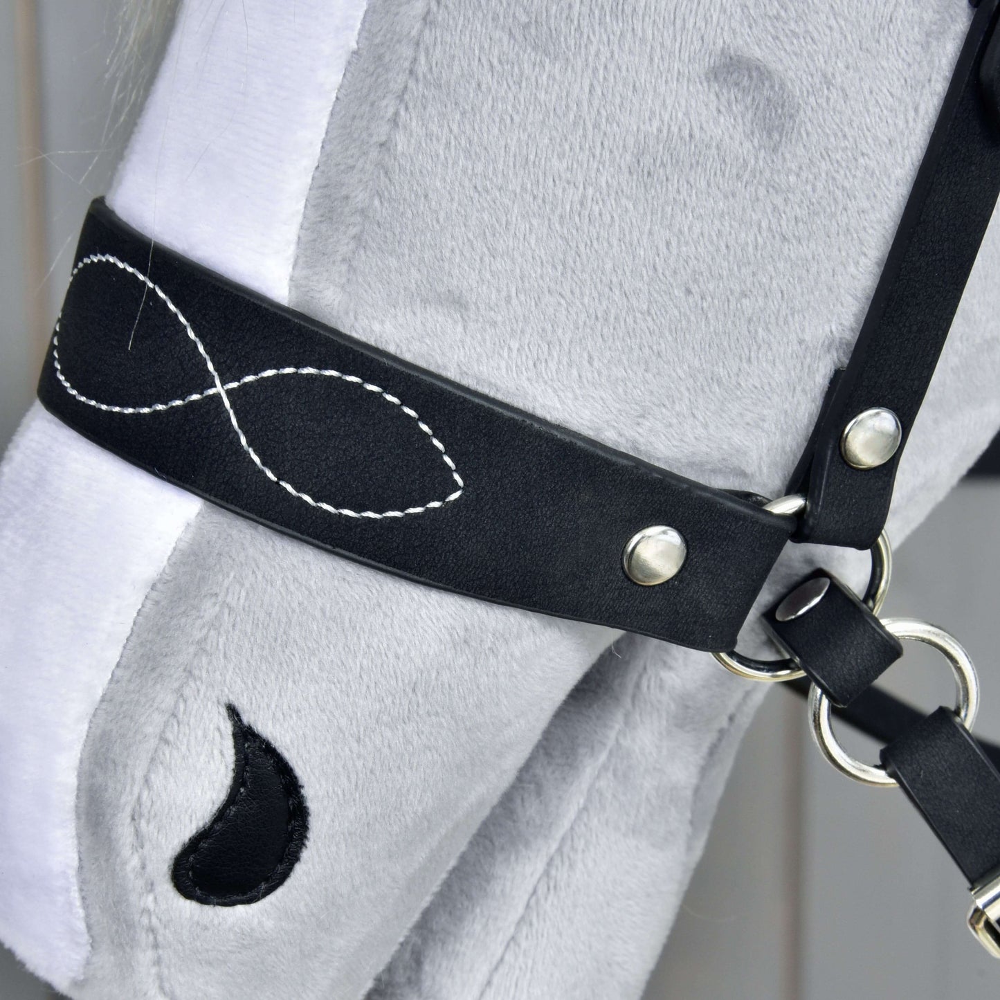 Detail hlavy šedého plyšového Hobby Horse s Kompletní uzdečkou bezudidlovou zdobenou od Hobby Horse LarDen, zdobenou bílým prošitím ve tvaru symbolu nekonečna a černou skvrnou ve tvaru kapky u oka.