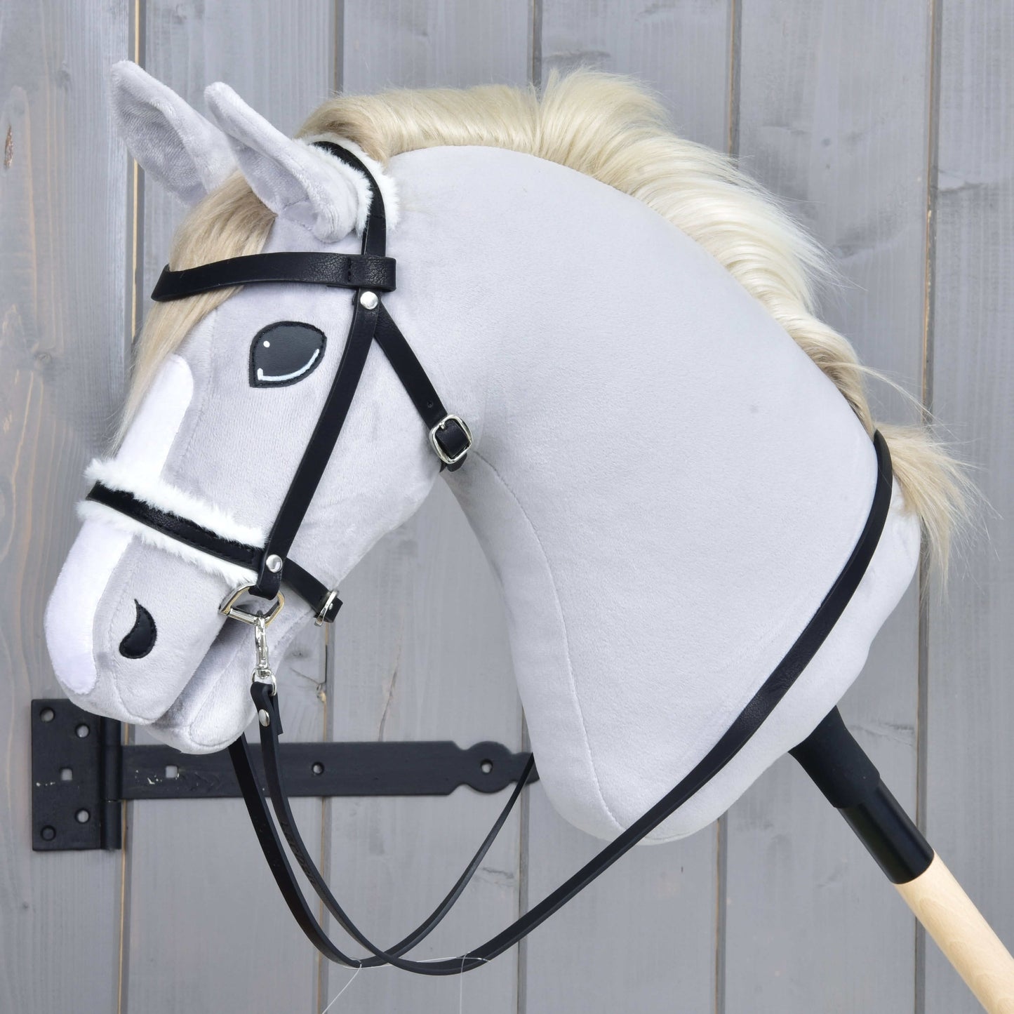 Kompletní uzdečka s beránkem černá od Hobby Horse LarDen na světle šedém Hobby Horse s krémovou hřívou, s černou koženkovou uzdečkou a otěžemi na dřevěné tyči, na pozadí šedého dřeva.