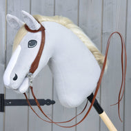 Plyšový Hobby Horse od Hobby Horse LarDen se světle šedou hlavou, bílou hřívou a Kompletní westernovou uzdečkou jednouška z kůže s dělenými otěžemi, upevněný na dřevěné tyči před šedou dřevěnou stěnou.