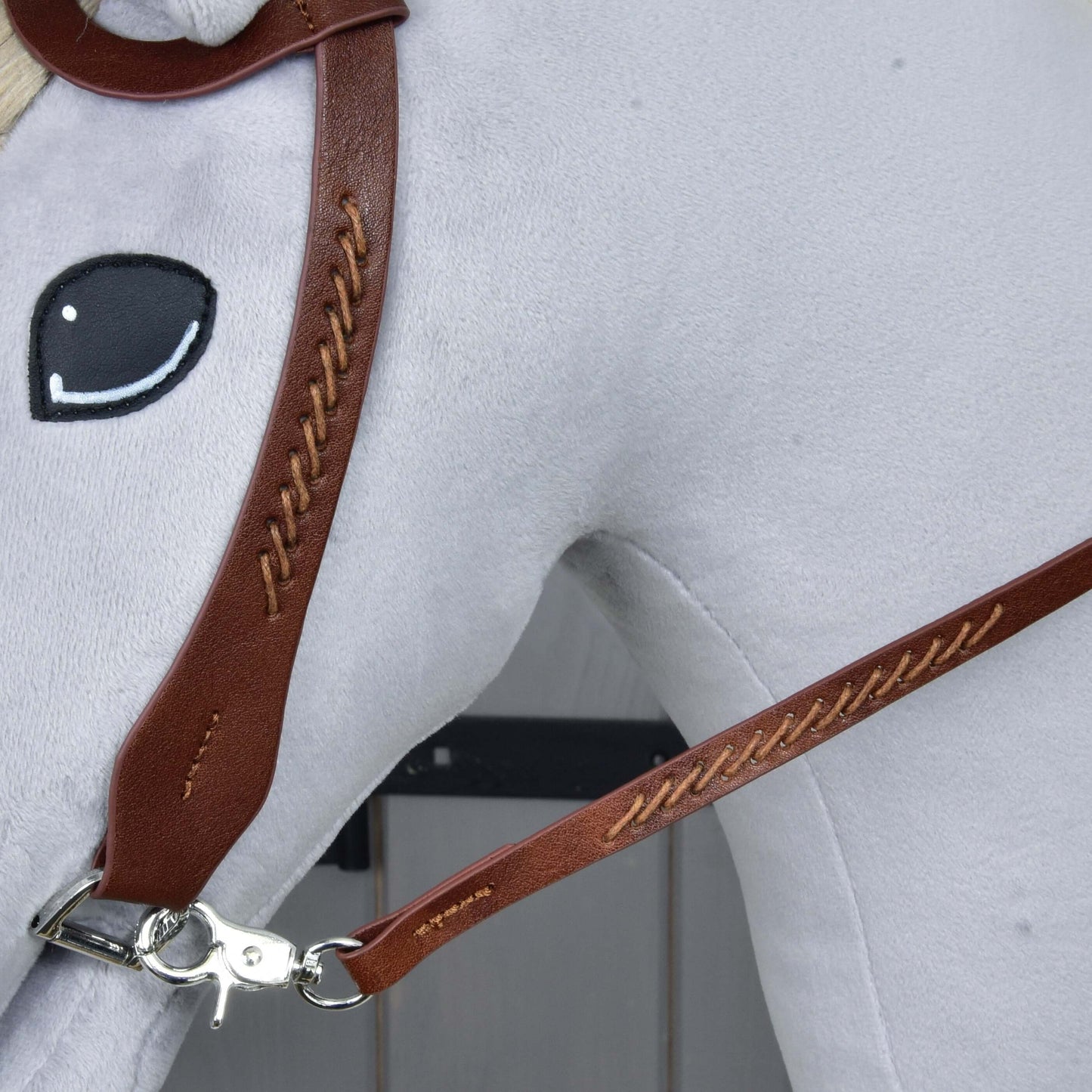 Detail plyšové koňské hlavy Hobby Horse LarDen s Kompletní westernovou uzdečkou jednouška z kůže z hnědé kůže, s výrazným prošíváním, dělenými otěžemi, kovovou sponou a jednoduchým černobíle vyšitým okem na světle šedém pozadí.