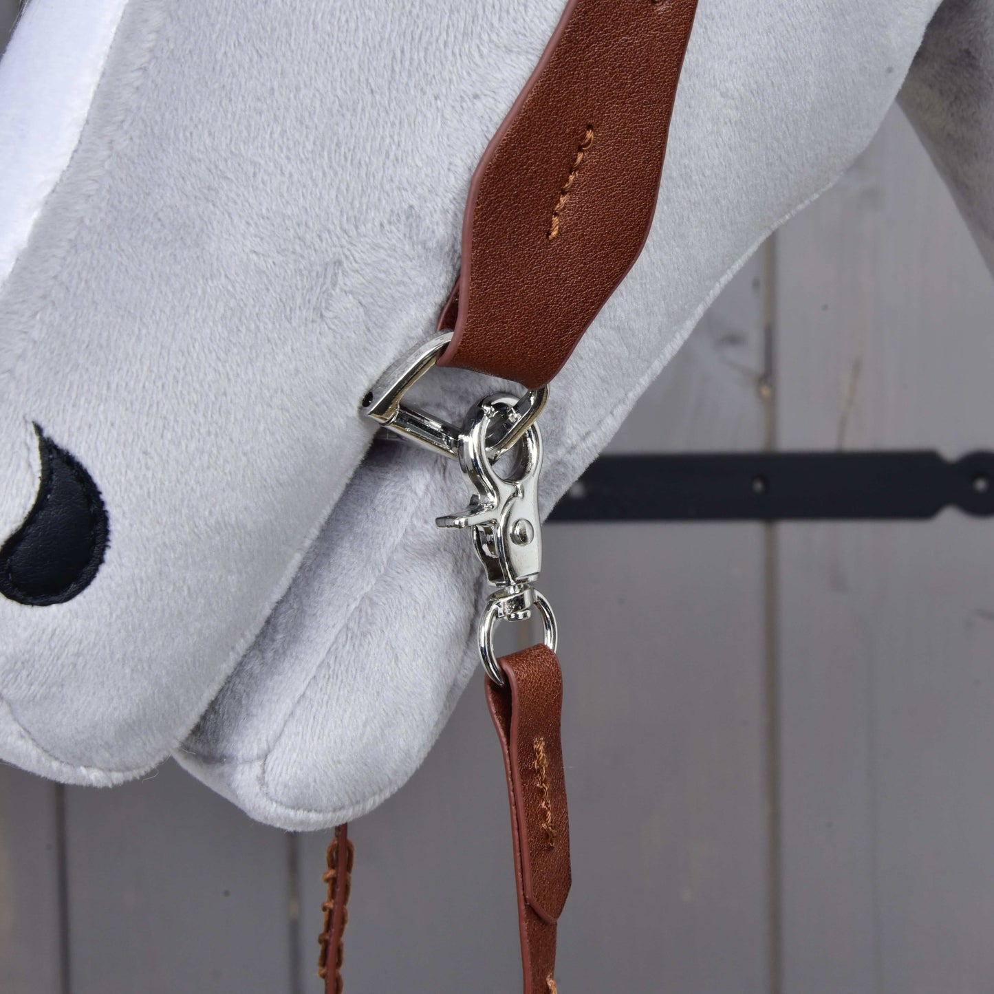 Detail Kompletní westernové uzdečky jednouška z kůže od Hobby Horse LarDen s hnědým koženým řemínkem, stříbrnou sponou a posuvným ušním poutkem, nasazené na hlavě bílého plyšového koně na pozadí šedého dřeva.