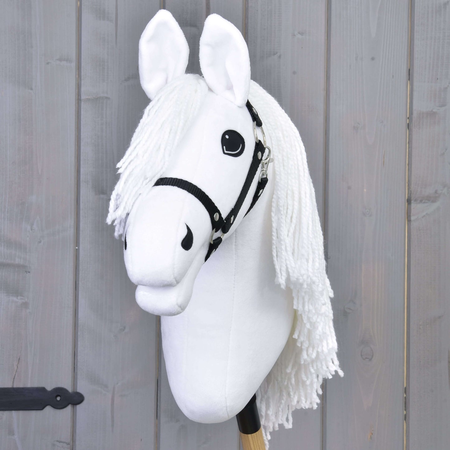 Hobby Horse Lady s černou ohlávkou od Hobby Horse LarDen s bílou hřívou a výraznýma očima na dřevěné tyči před šedým prkenným pozadím, ideální pro nápaditou drezurní hru inspirovanou lipicány.