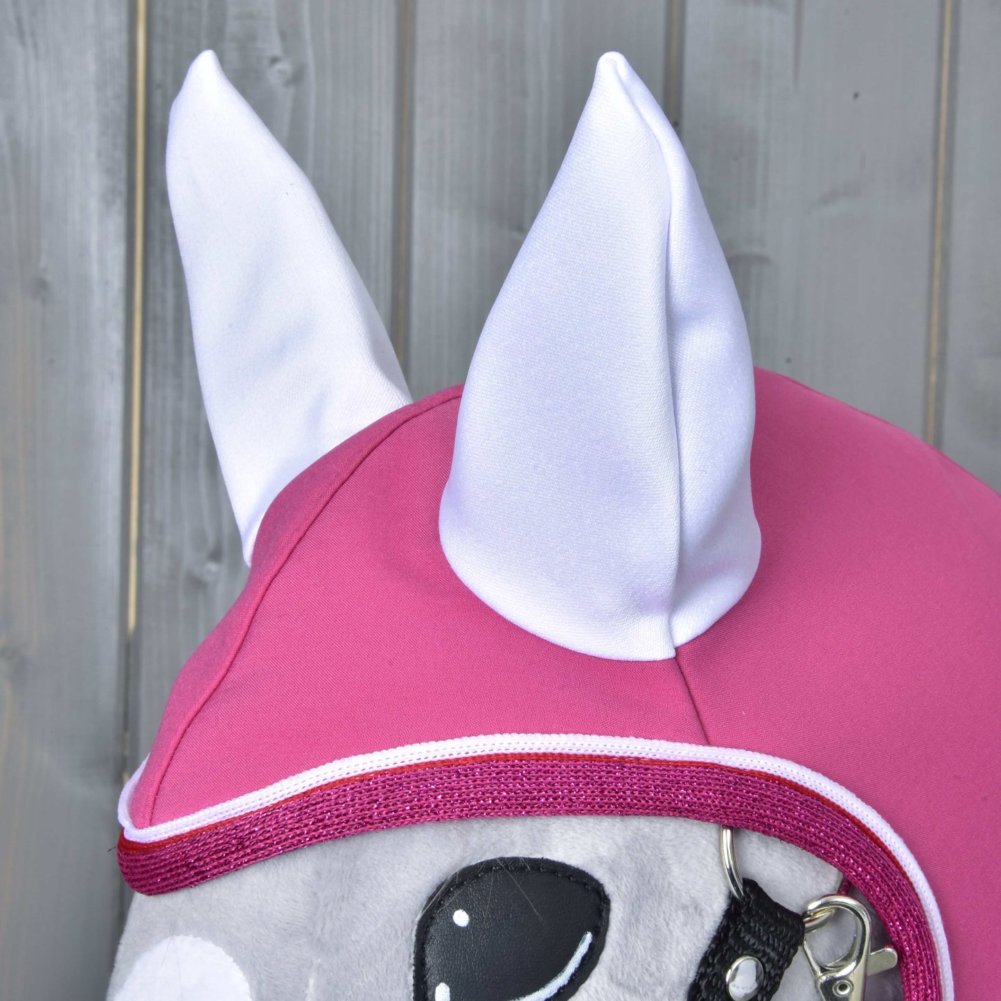 Detail Pláštěnky LarDen od Hobby Horse LarDen na plyšové hračce koně s bílýma látkovýma ušima a jasně růžovými doplňky uzdečky na světle šedém dřevěném pozadí.