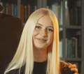 Mladá žena s dlouhými rovnými blond vlasy se usmívá do kamery. Má na sobě černý top a za ní je rozmazaná knihovna. Ruka jí jemně spočívá na rameni, což naznačuje vřelý okamžik po zábavném dni s hobby horsing.
