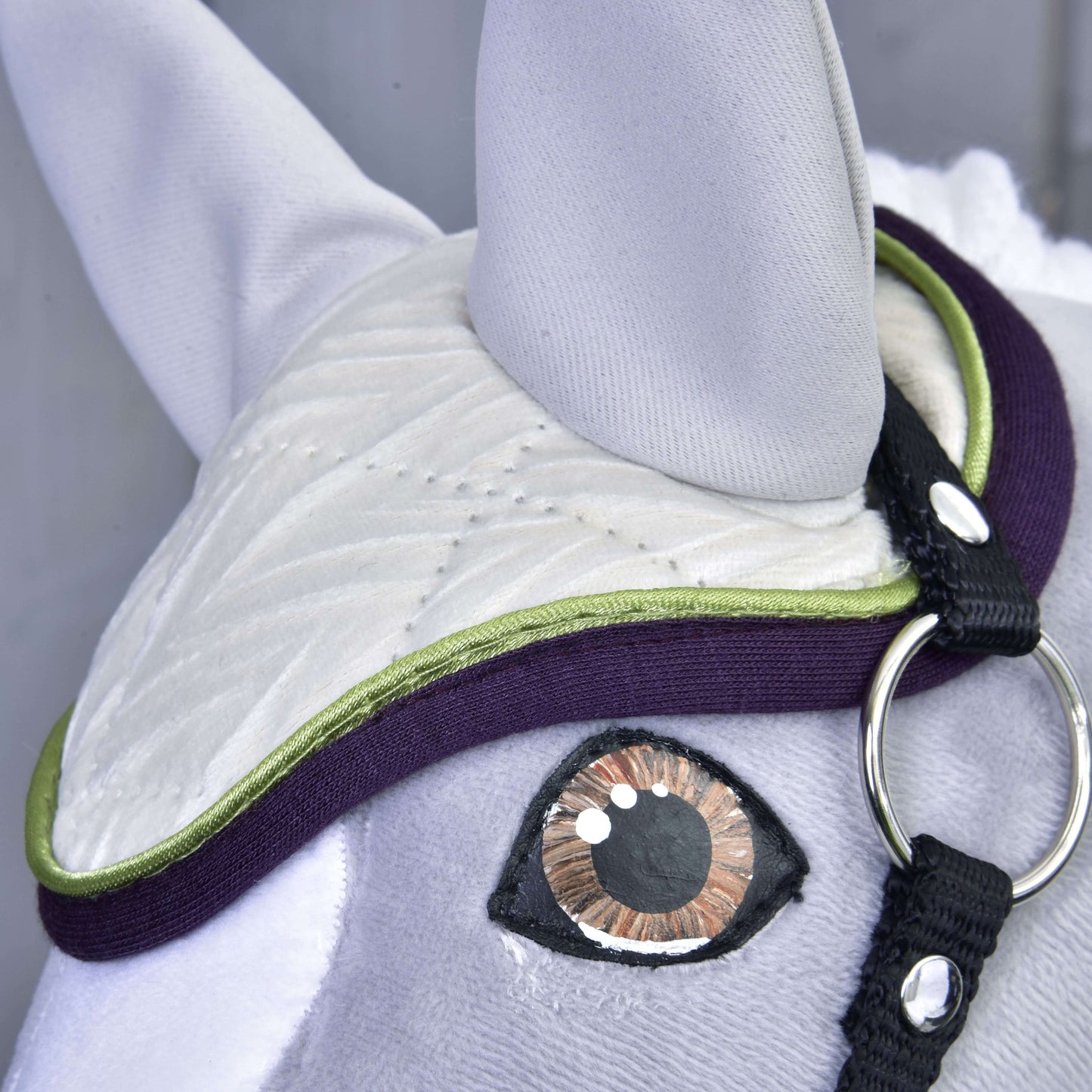 Detail Set Leonardo od Hobby Horse LarDen: látková koňská hlava s prošívanými detaily, velkým hnědo-černým okem, bílou prošívanou čabrakou se zeleným a fialovým lemováním, černým popruhem a stříbrným kroužkem – půvabná dekorace.