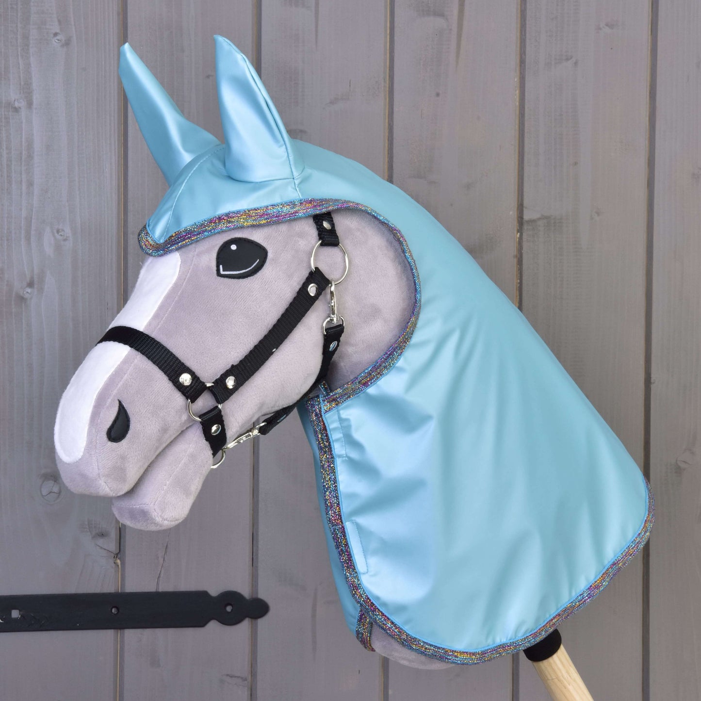 Šedý Hobby Horse od Hobby Horse LarDen s černou uzdečkou, velkýma plstěnýma očima a kapucí Pláštěnka Light Blue s ladícíma ušima, zobrazený před dřevěnou stěnou.