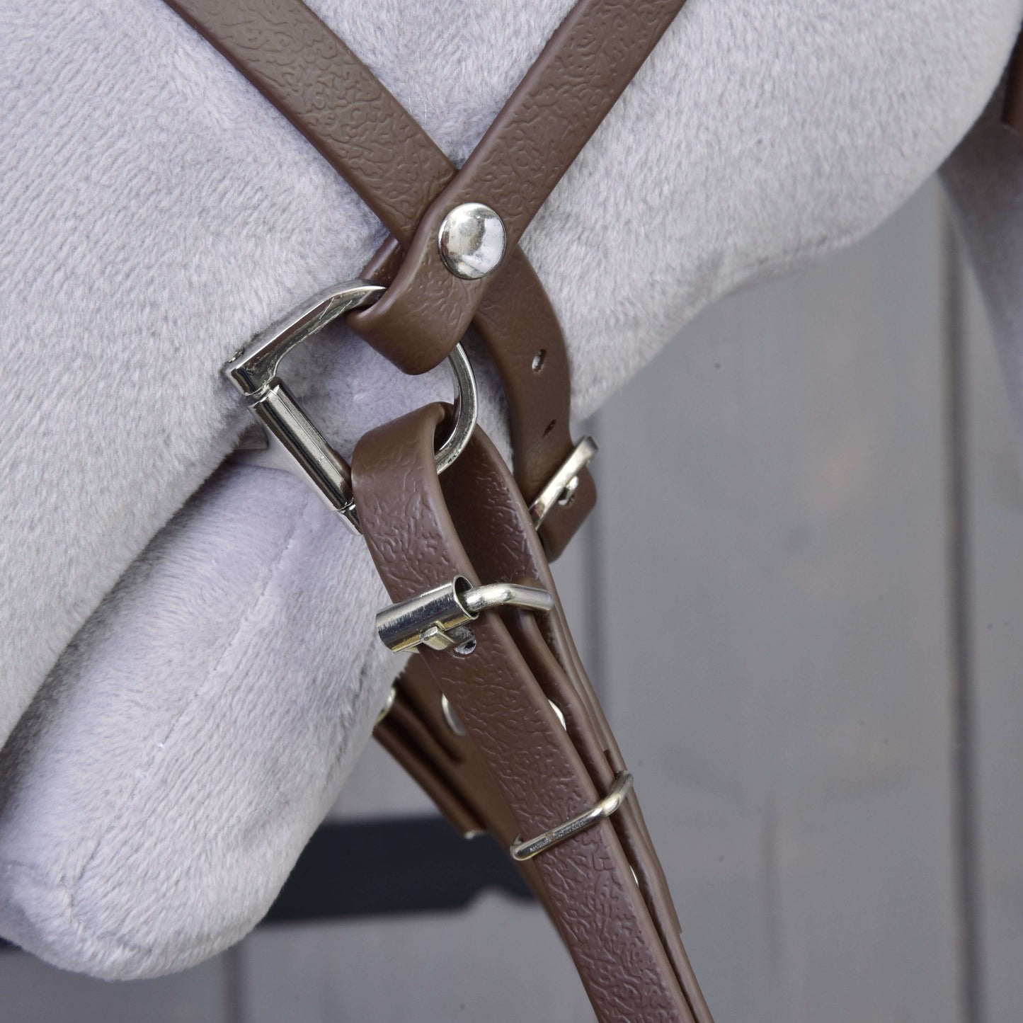 Detail Kompletní uzdečka s nánosníkem Brown od Hobby Horse LarDen v hnědé barvě s kovovou přezkou, připevněné ke světle šedému povrchu, s rozmazaným dřevěným pozadím.