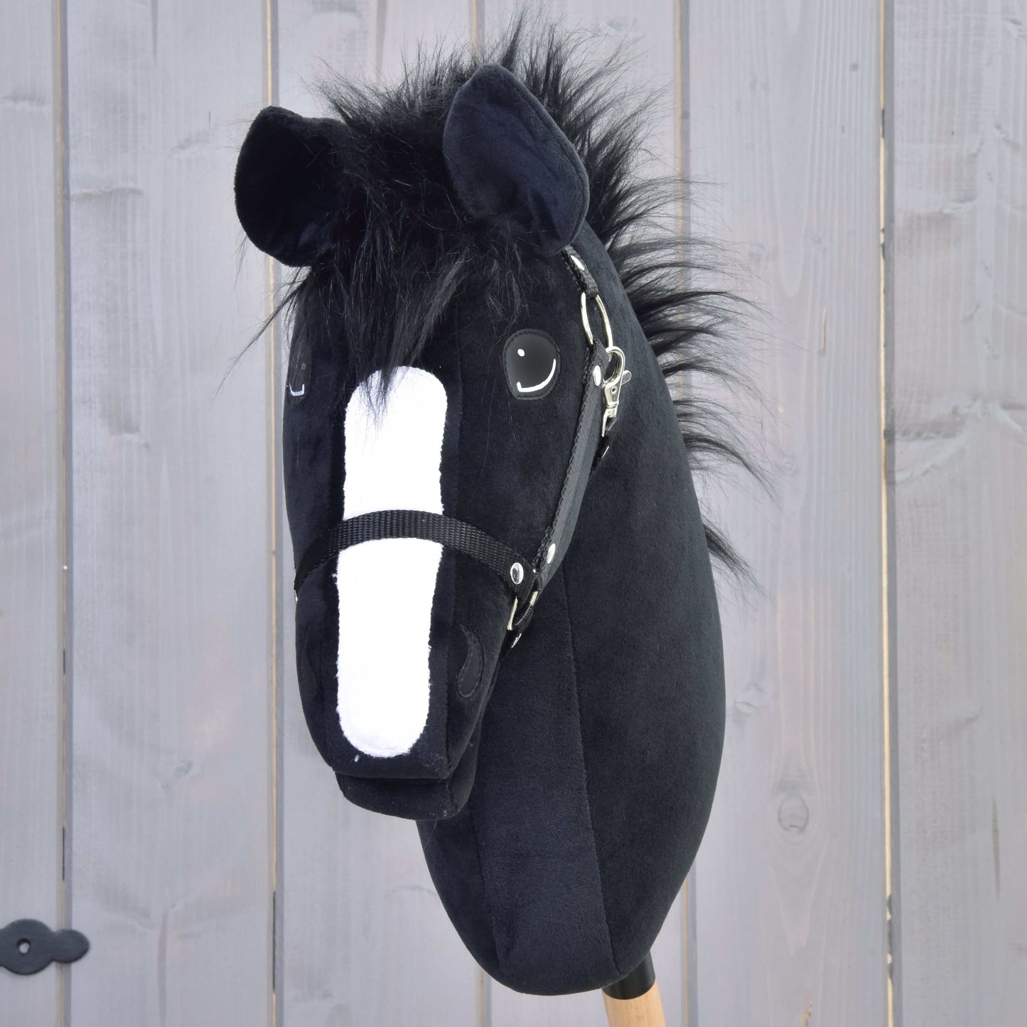 Hobby Horse Loki s černou ohlávkou od Hobby Horse LarDen má černé plyšové provedení ve stylu Welsh Coba, bílý čenich, huňatou hřívu a dřevěnou tyč – ideální pro drezurní hru před světle šedým plotem.