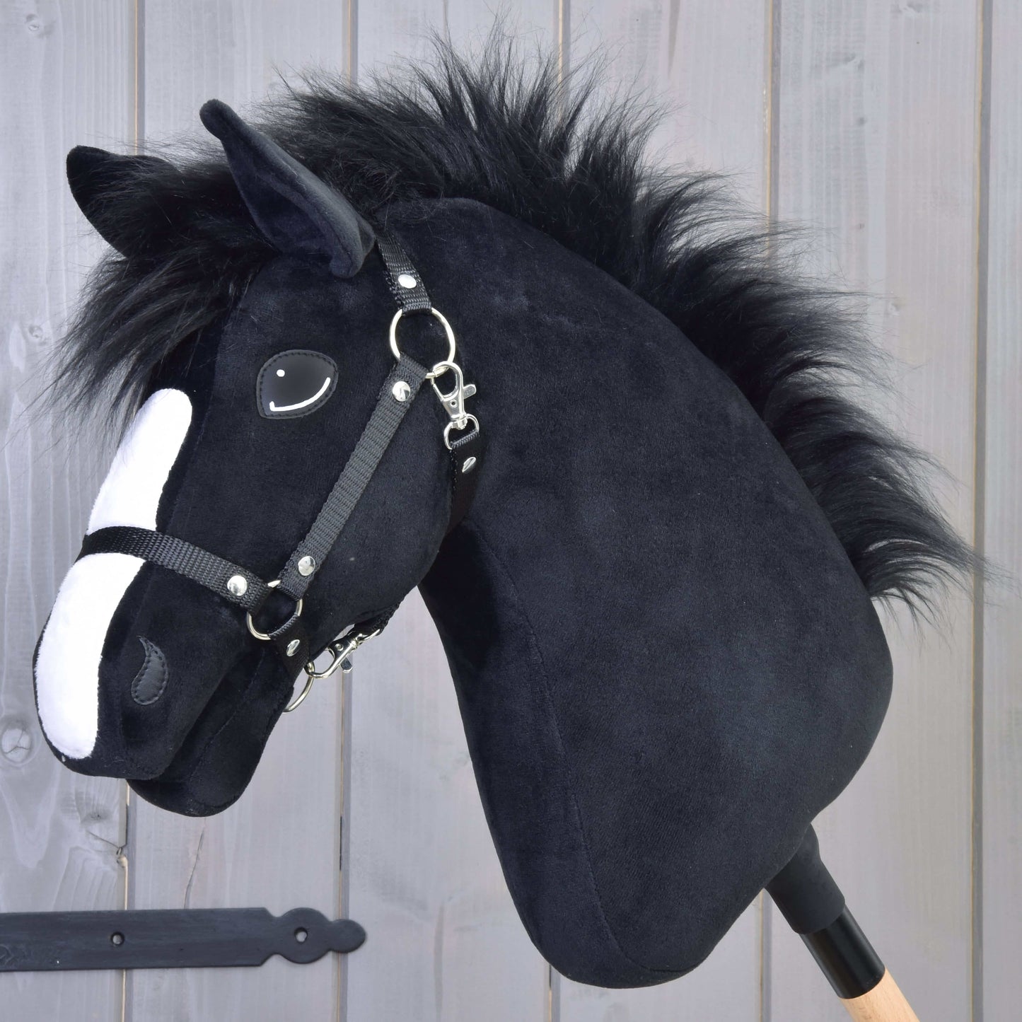 Hobby Horse Loki s černou ohlávkou od Hobby Horse LarDen má černé plyšové tělo, bílý pruh na hlavě a černou ohlávku. Je upevněný na dřevěné tyči a zasazený před světlý dřevěný panel – ideální pro hru v drezurním stylu.
