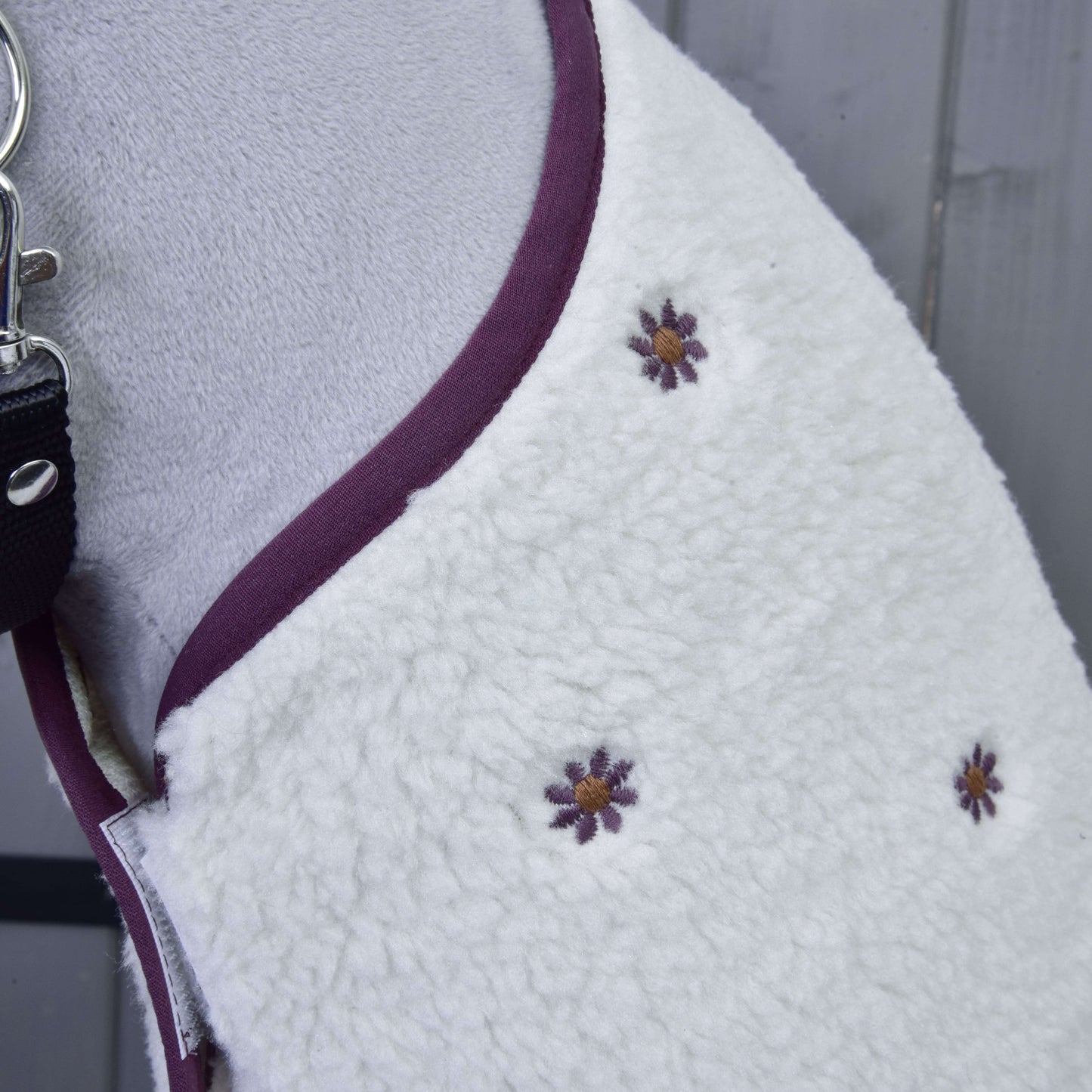 Detail Set Loret, bílého fleecového psího kabátku od Hobby Horse LarDen, s fialovo-žlutě vyšitými květy a fialovým lemem. Zobrazený na psovi s černým vodítkem připomíná deku pro Hobby Horse a leží na šedém dřevěném povrchu.