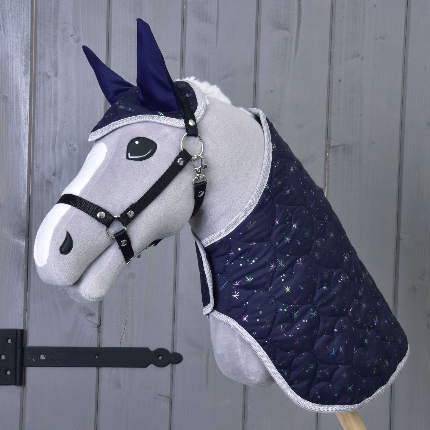 Hobby Horse LarDen Set Magic obsahuje plyšového Hobby Horse s tmavě modrou prošívanou dekou a náušníkem se vzorem hvězd, poskytující ochranu proti hmyzu, zobrazeného před dřevěnou stěnou.
