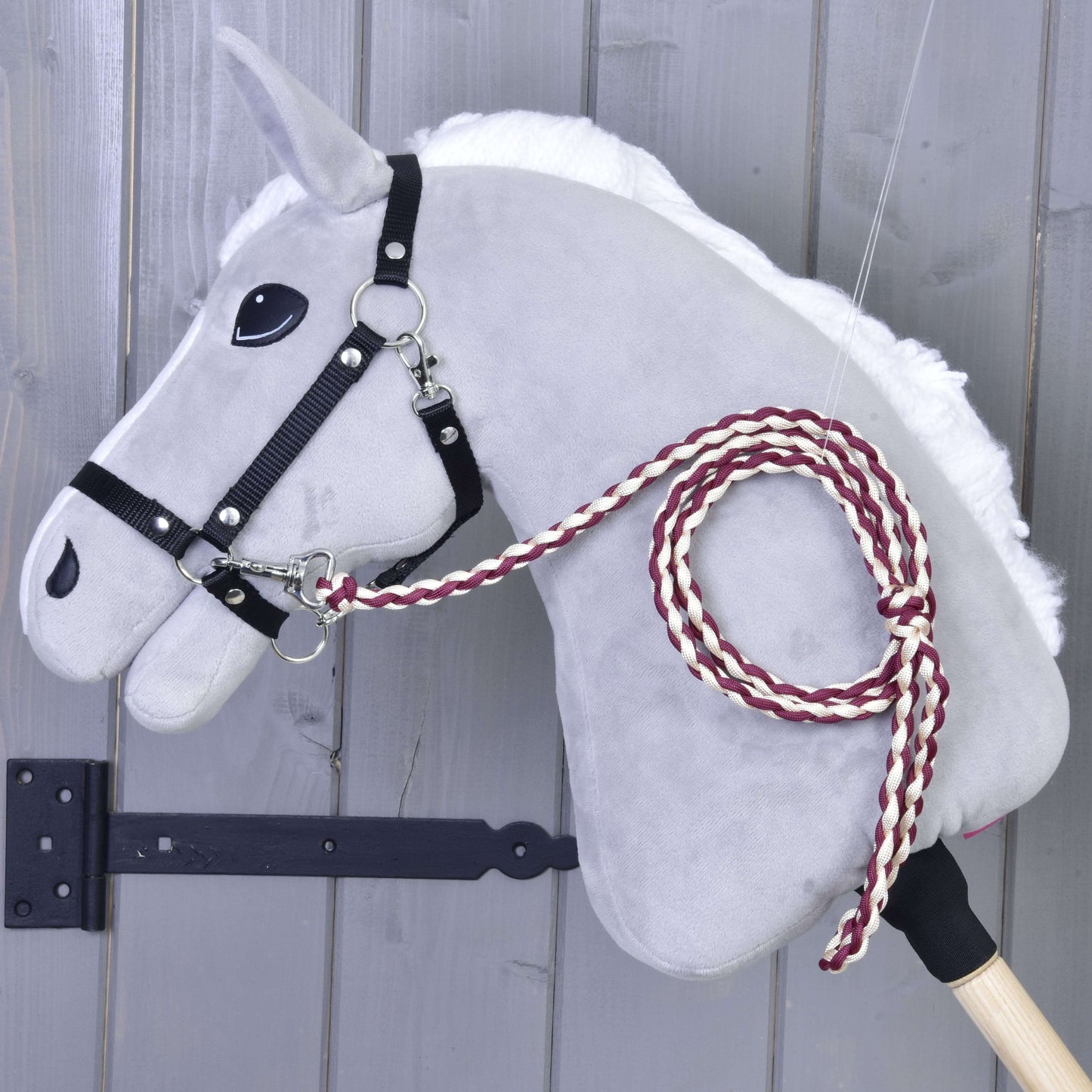 Hobby Horse - pletené vodítko Maroon od Hobby Horse LarDen se šedou plstěnou hlavou, černou uzdečkou a ručně pleteným vínovo-bílým vodítkem, nasazené na dřevěné tyči před šedou dřevěnou stěnou.