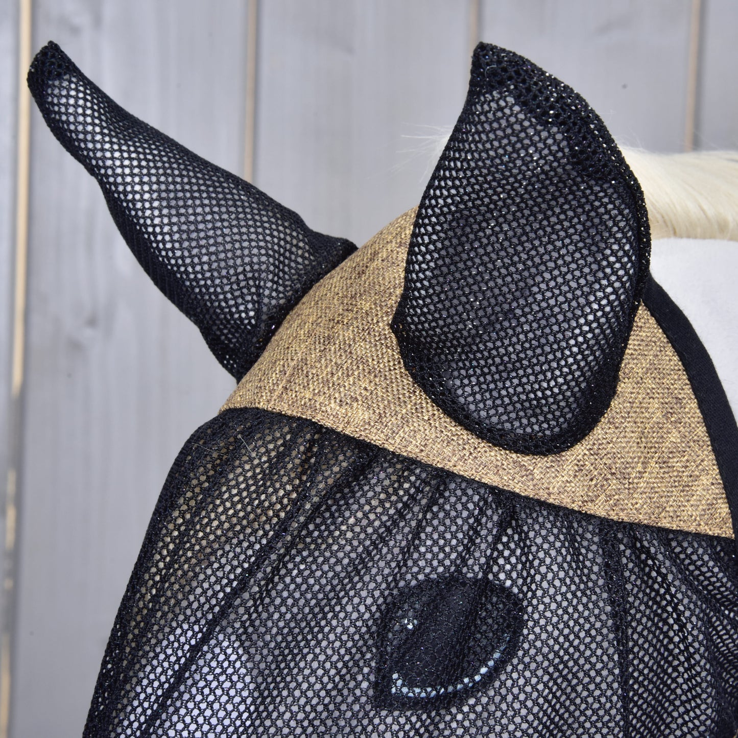 Detail koně s maskou proti hmyzu de Luxe Mona od Hobby Horse LarDen. Tato černá síťovaná maska s kryty na uši chrání koňské uši a oči před hmyzem – ideální pro skutečné koně i fanoušky Hobby Horse. Světlé dřevěné pozadí.