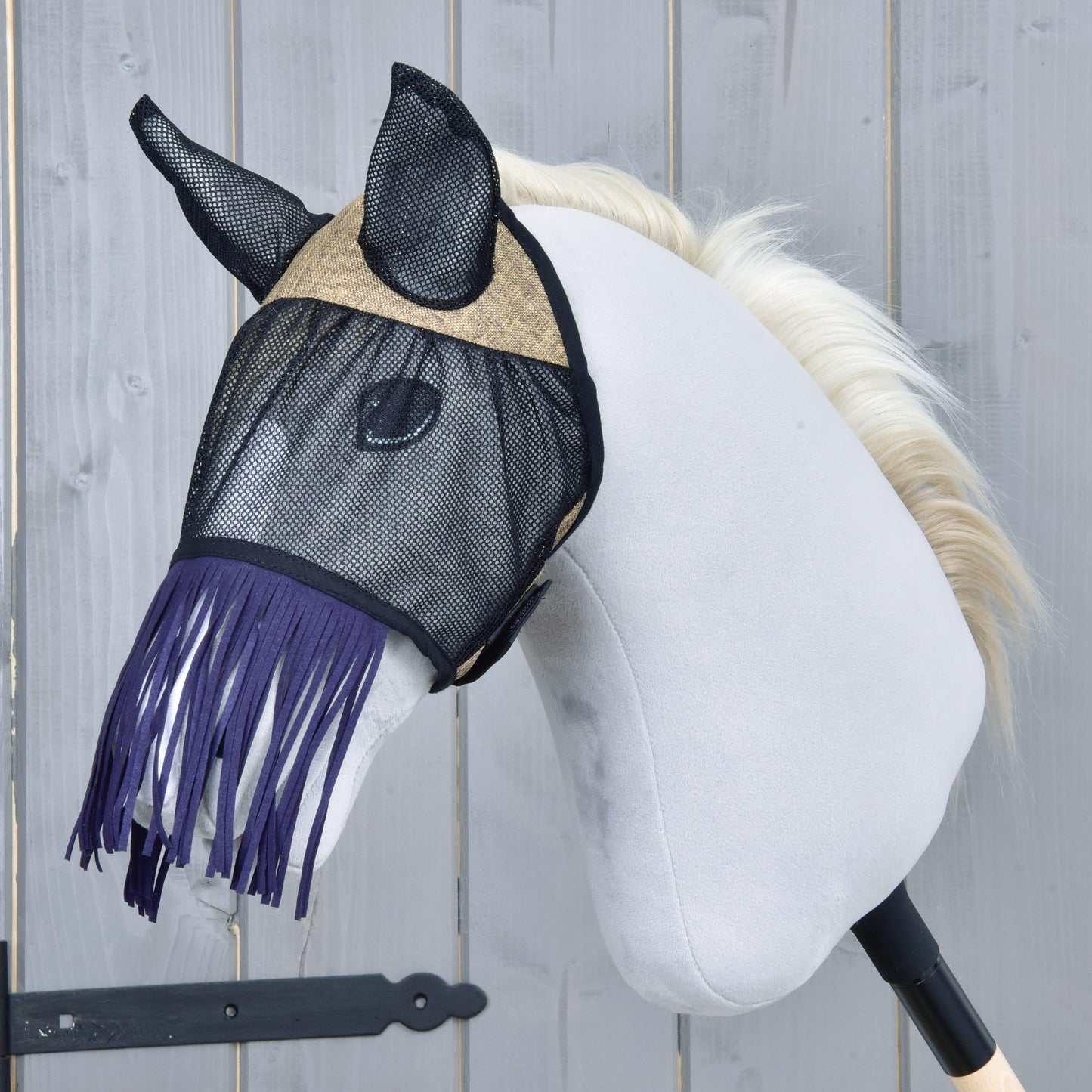 Maska proti hmyzu de Luxe Mona od Hobby Horse LarDen na bílém plyšovém Hobby Horse s černou tyčí a světle blond hřívou, se stylovou třásňovou maskou proti hmyzu na pozadí šedé dřevěné stěny.