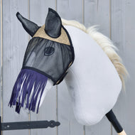 Maska proti hmyzu de Luxe Mona od Hobby Horse LarDen na bílém plyšovém Hobby Horse s černou tyčí a světle blond hřívou, se stylovou třásňovou maskou proti hmyzu na pozadí šedé dřevěné stěny.