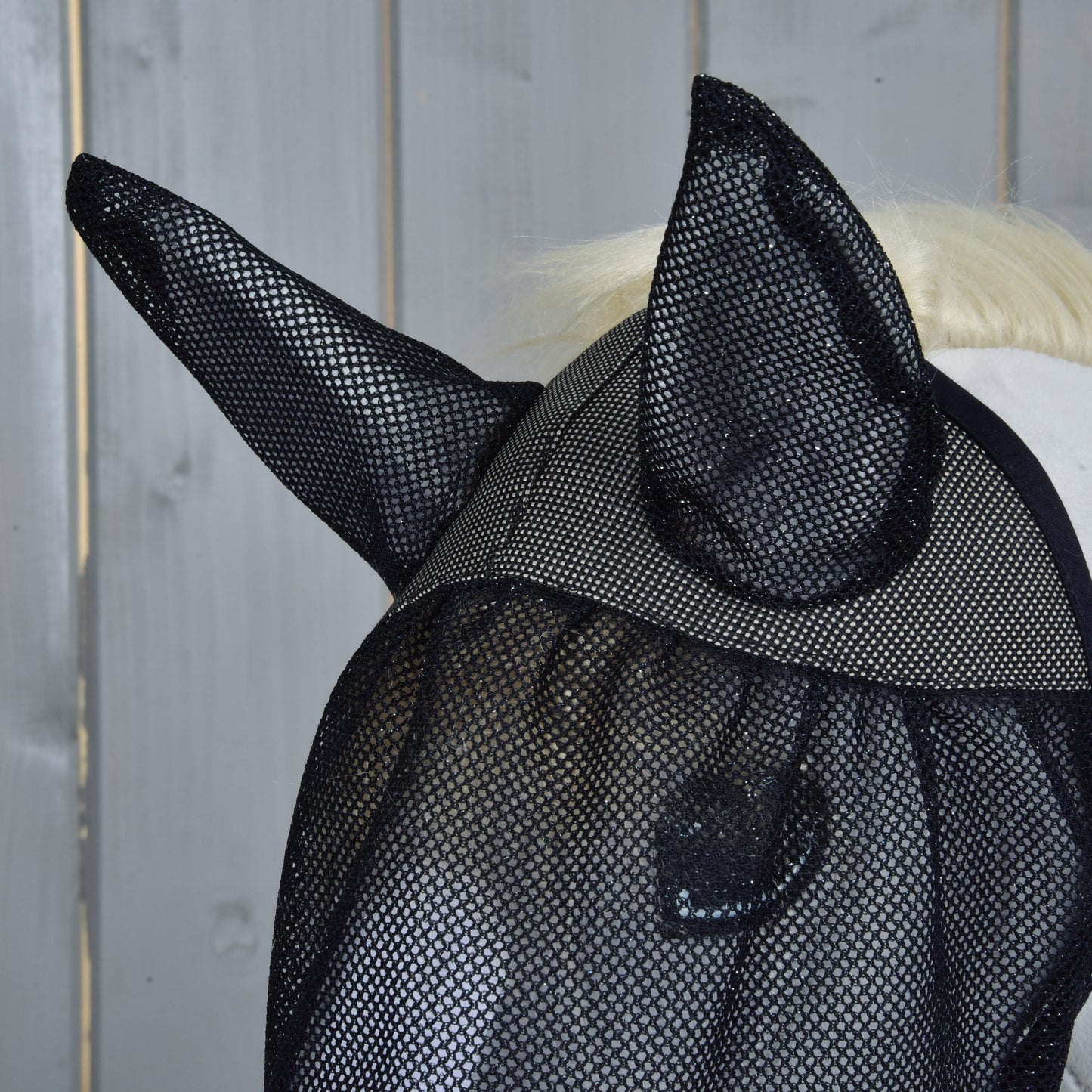 Detail koně s maskou proti hmyzu de Luxe Full od Hobby Horse LarDen s kryty uší, před šedým dřevěným pozadím.