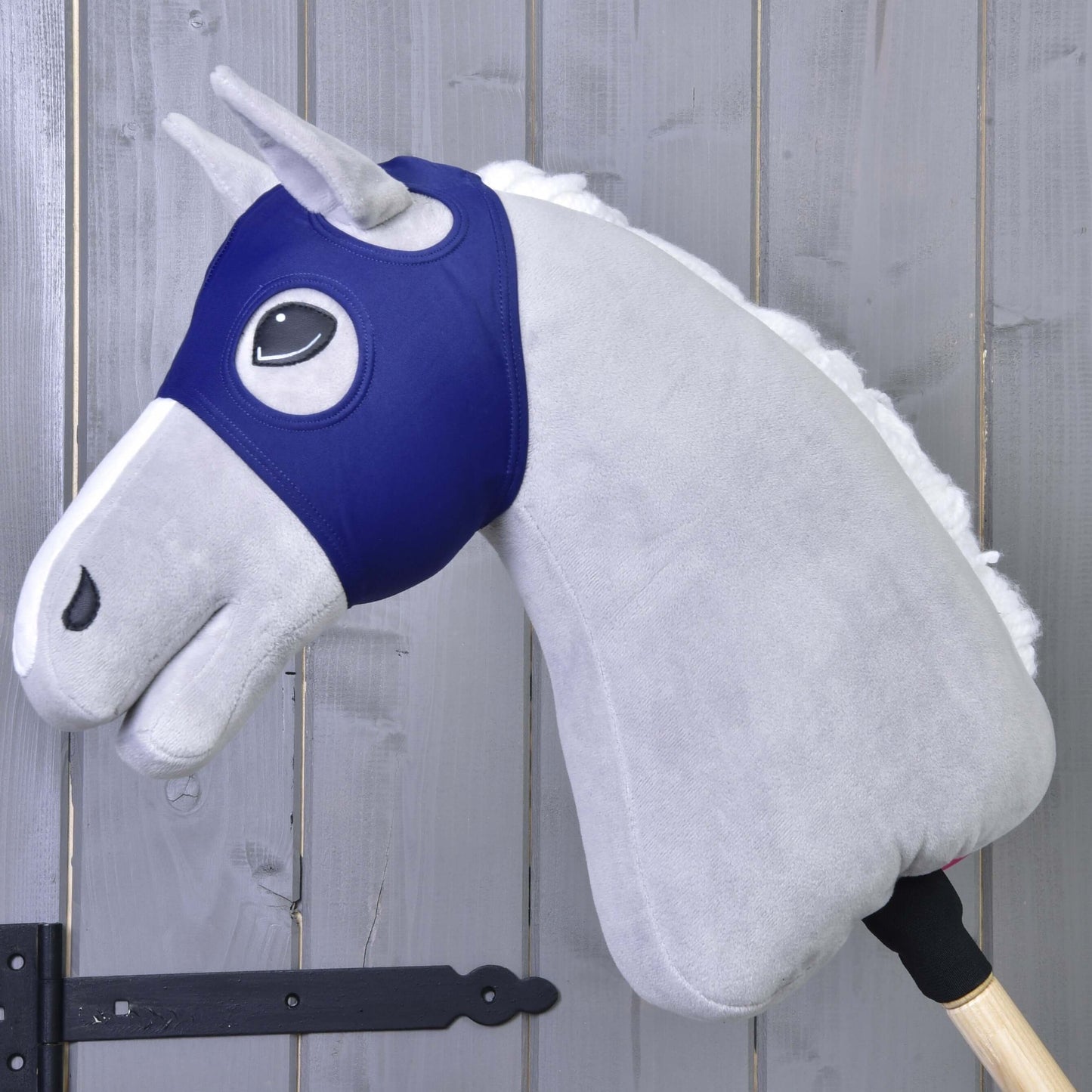 Šedý Hobby Horse LarDen s bílou hřívou a dřevěnou tyčí je zobrazený s maskou bez uší LarDen před dřevěnou stěnou s viditelnými panty.