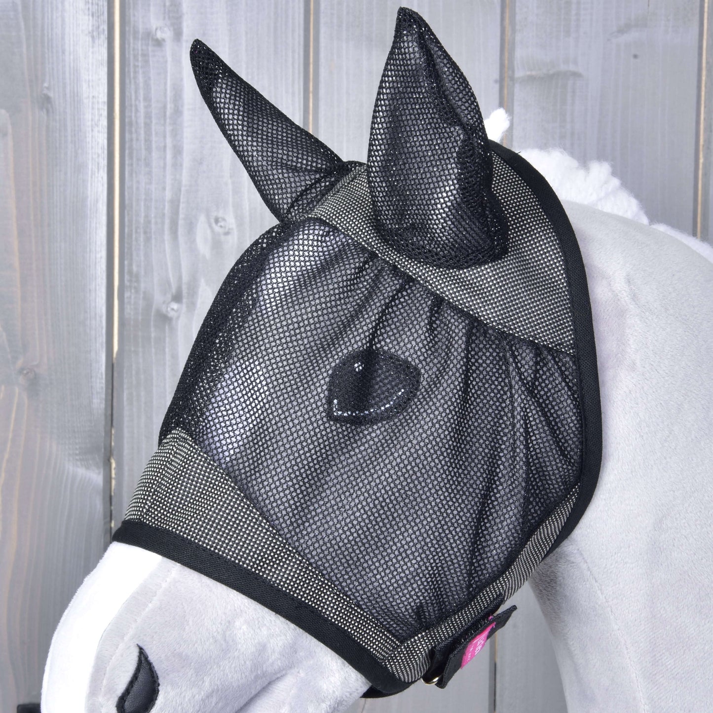 Bílý kůň stojí u dřevěného plotu a má na sobě Hobby Horse LarDen Maska proti hmyzu de Luxe Grey, omyvatelnou masku s kryty uší, která chrání jeho oči, uši a část obličeje před hmyzem.
