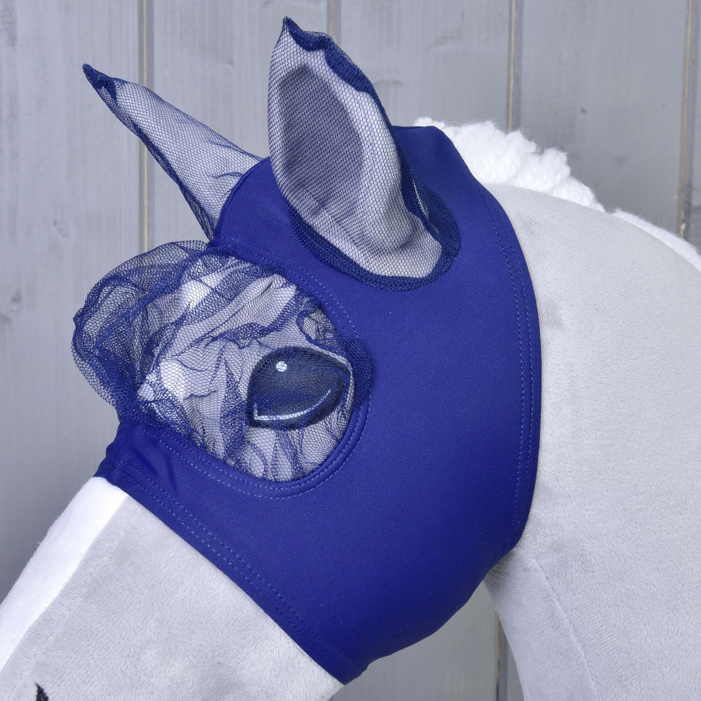 Bílá plyšová hračka Hobby Horse LarDen předvádí modrou pratelnou masku proti hmyzu Fly Mask LarDen, která zakrývá uši a oči pro ochranu, na světlém dřevěném pozadí.
