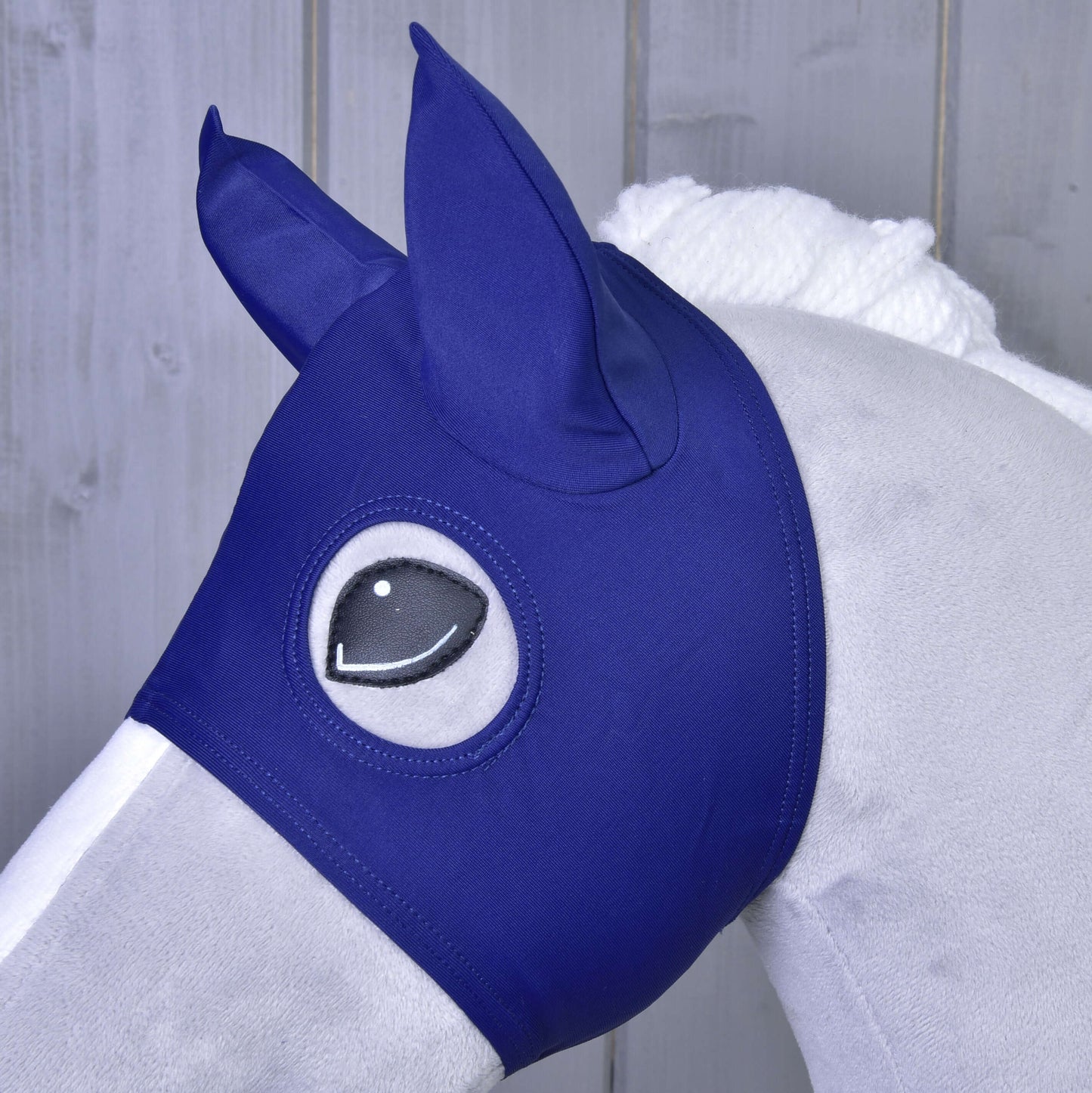 Detail šedého koně s maskou Maska s ušima LarDen od Hobby Horse LarDen, s kryty uší a otvory pro oči, stojícího před dřevěným pozadím.