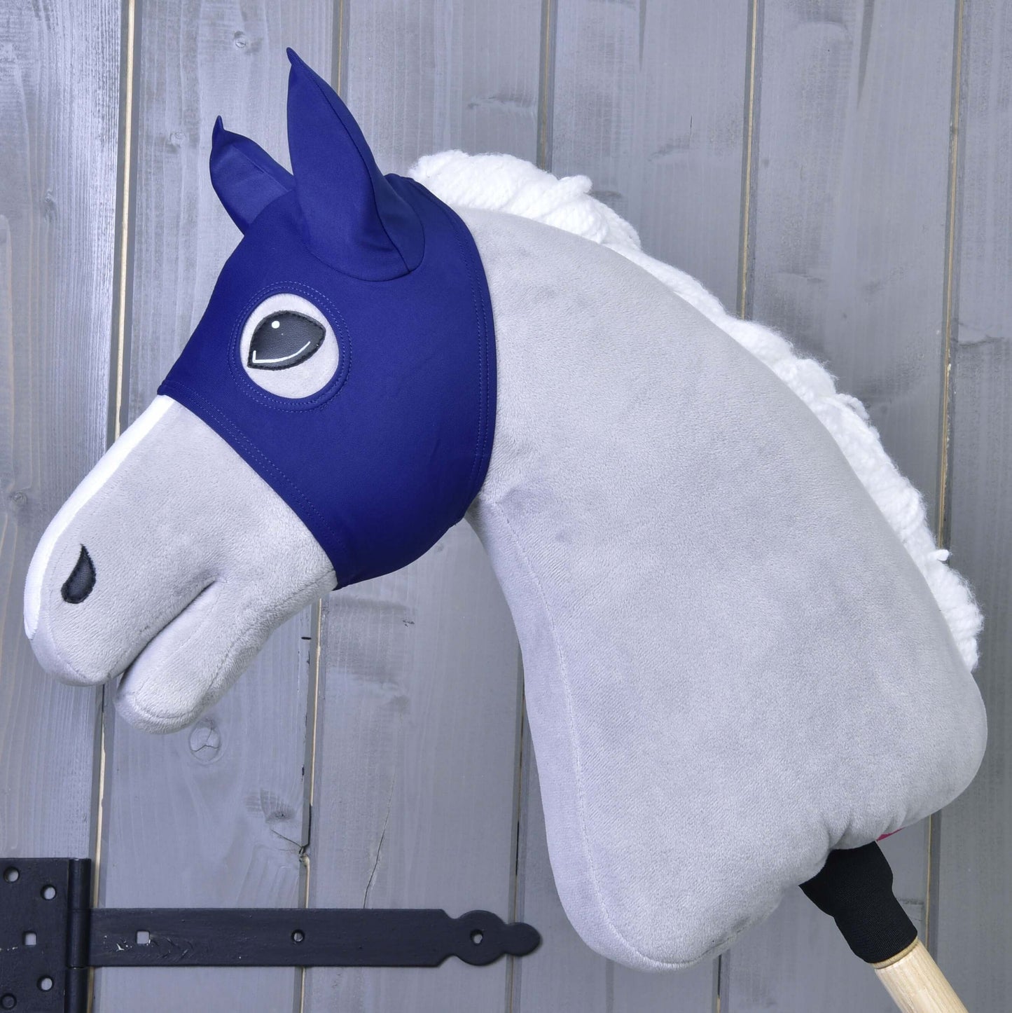 Maska s ušima LarDen od Hobby Horse LarDen na šedém plyšovém Hobby Horse s modrou látkovou uzdečkou a bílou hřívou na dřevěné tyči – ideální pro nápaditou hru PES, zobrazená před šedou dřevěnou stěnou.