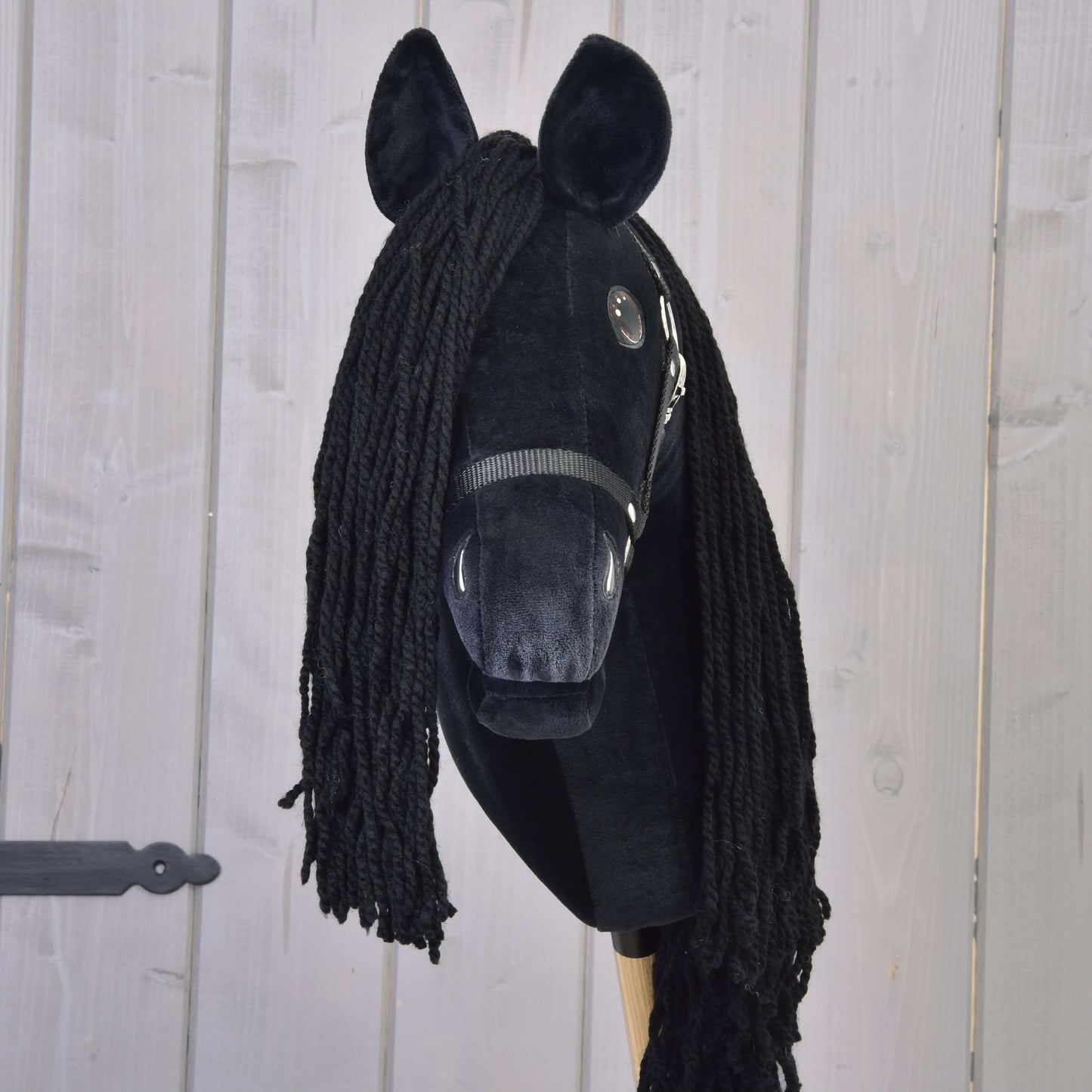 Hobby Horse Maximus s černou ohlávkou od Hobby Horse LarDen, ručně vyráběný fríský Hobby Horse s plyšovou hlavou, přízovou hřívou, knoflíkovými oči, černou ohlávkou a dřevěnou tyčí, zobrazený na světlém dřevěném pozadí.