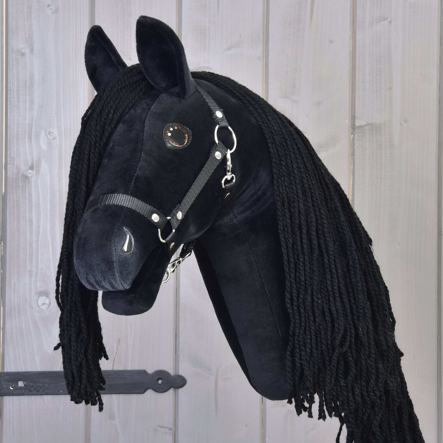 Hobby Horse Maximus s černou ohlávkou od Hobby Horse LarDen, plyšový černý fríský Hobby Horse s dlouhou černou hřívou, na dřevěné tyči, zobrazený před světlým dřevěným obložením.
