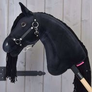 Hobby Horse Maximus s černou ohlávkou od Hobby Horse LarDen s lesklýma knoflíkovýma očima a přízovou hřívou, na dřevěné tyči, před stěnou ze světlých dřevěných prken.