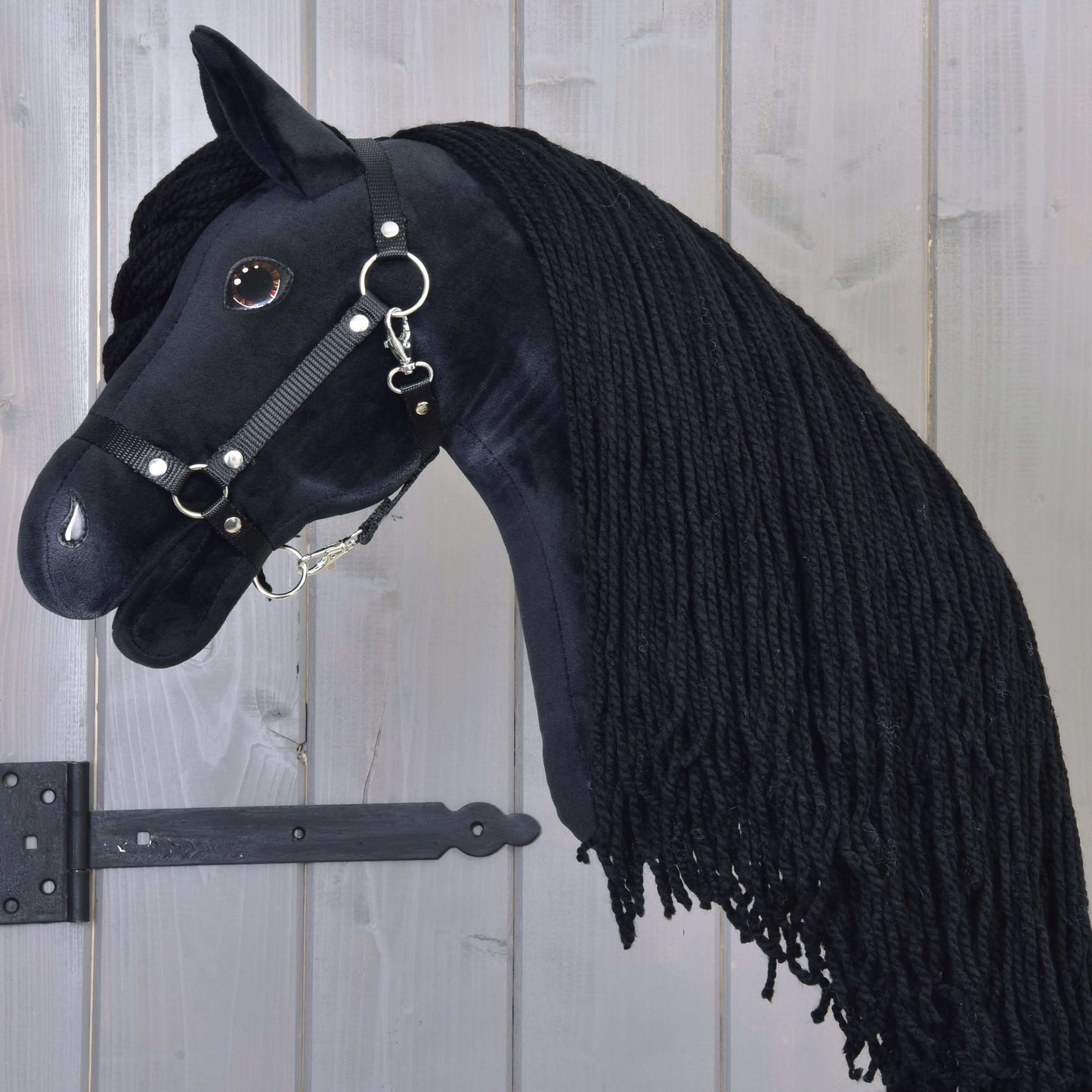 Hobby Horse Maximus s černou ohlávkou od Hobby Horse LarDen s plyšovým černým fríským vzhledem, dlouhou zapletenou hřívou, uzdečkou s kovovými kroužky a lesklým knoflíkovým okem, zavěšený na dřevěné stěně.