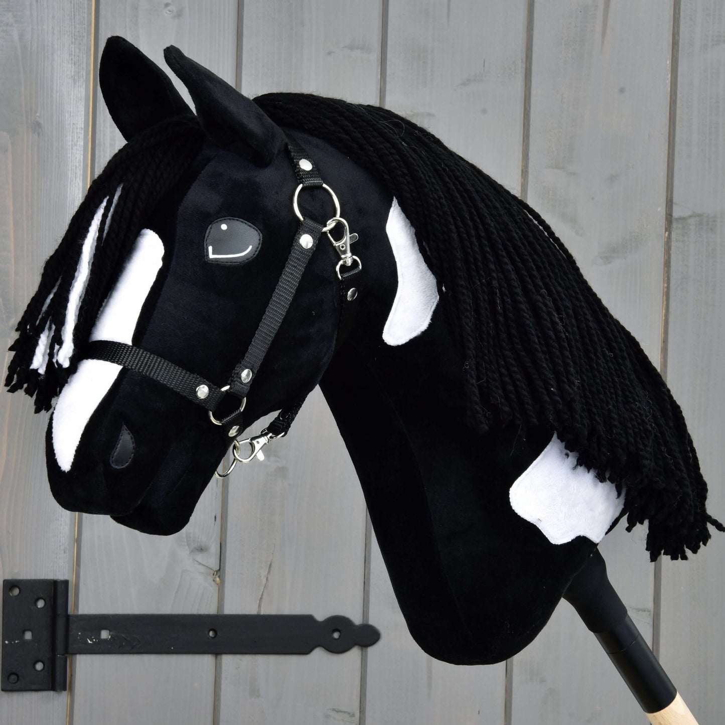 Hobby Horse Merlin s černou ohlávkou od Hobby Horse LarDen, inspirovaný hřebcem plemene Barock Pinto, má černobílé skvrny a zapletenou přízovou hřívu, upevněný před šedou dřevěnou stěnou.