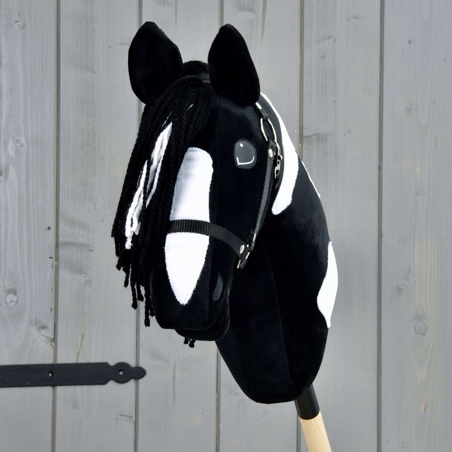Hobby Horse Merlin s černou ohlávkou od Hobby Horse LarDen je černobíle skvrnitý Hobby Horse ve stylu hřebce Barock Pinto, s přízovou hřívou a uzdečkou, upevněný na dřevěné tyči před šedým plotem.