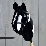 Hobby Horse Merlin s černou ohlávkou od Hobby Horse LarDen je černobíle skvrnitý Hobby Horse ve stylu hřebce Barock Pinto, s přízovou hřívou a uzdečkou, upevněný na dřevěné tyči před šedým plotem.