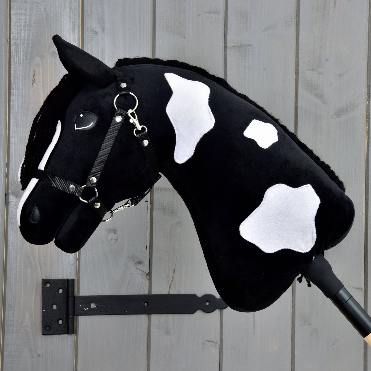 Hobby Horse Merlin s černou ohlávkou od Hobby Horse LarDen, černobílý plyšový hřebec Barock Pinto s usměvavou tváří a černou hřívou, je zobrazený upevněný na dřevěné stěně.