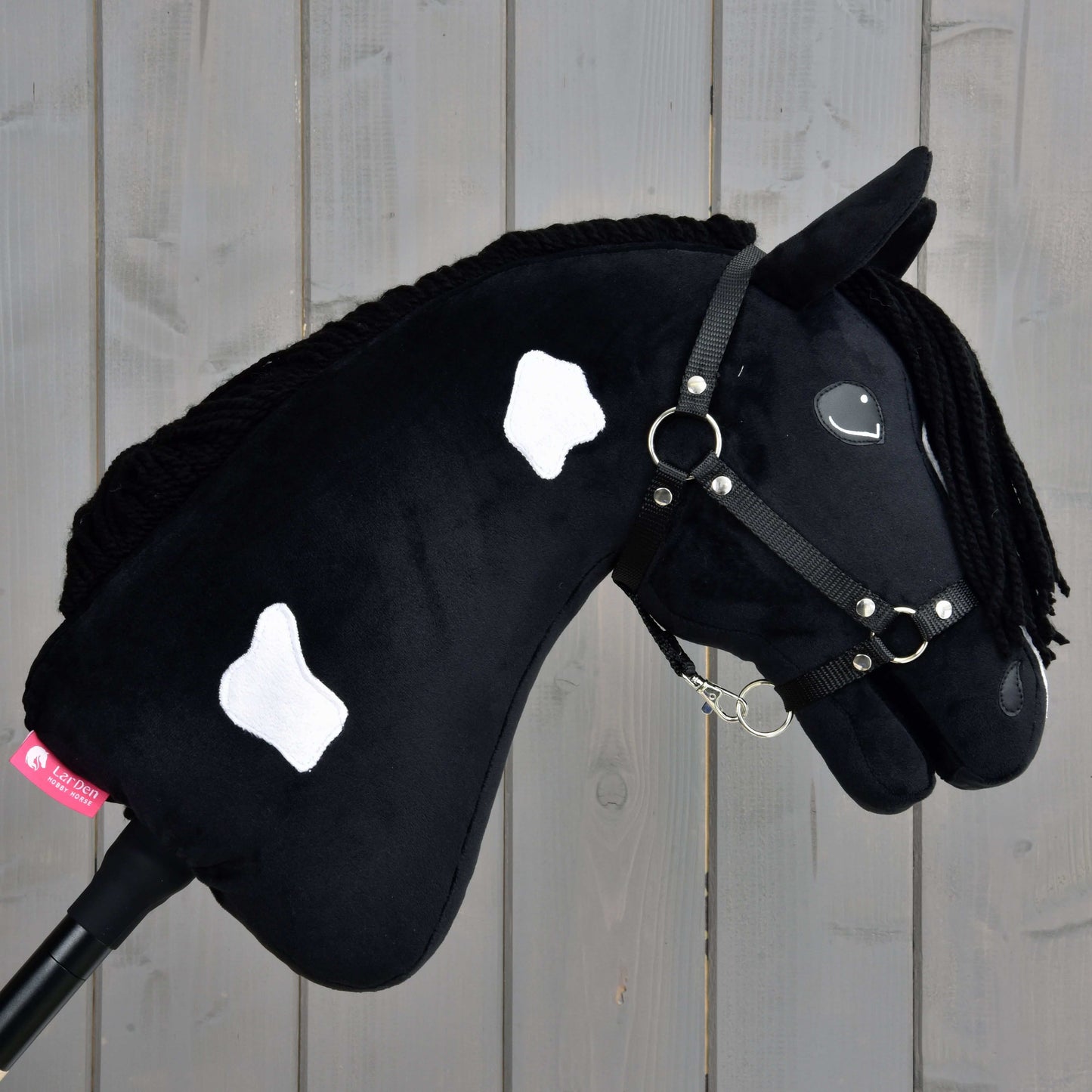 Hobby Horse Merlin s černou ohlávkou od Hobby Horse LarDen má černé tělo inspirované Barock Pinto s bílými skvrnami, zapletenou hřívu a černou ohlávku, na pozadí dřevěných prken.
